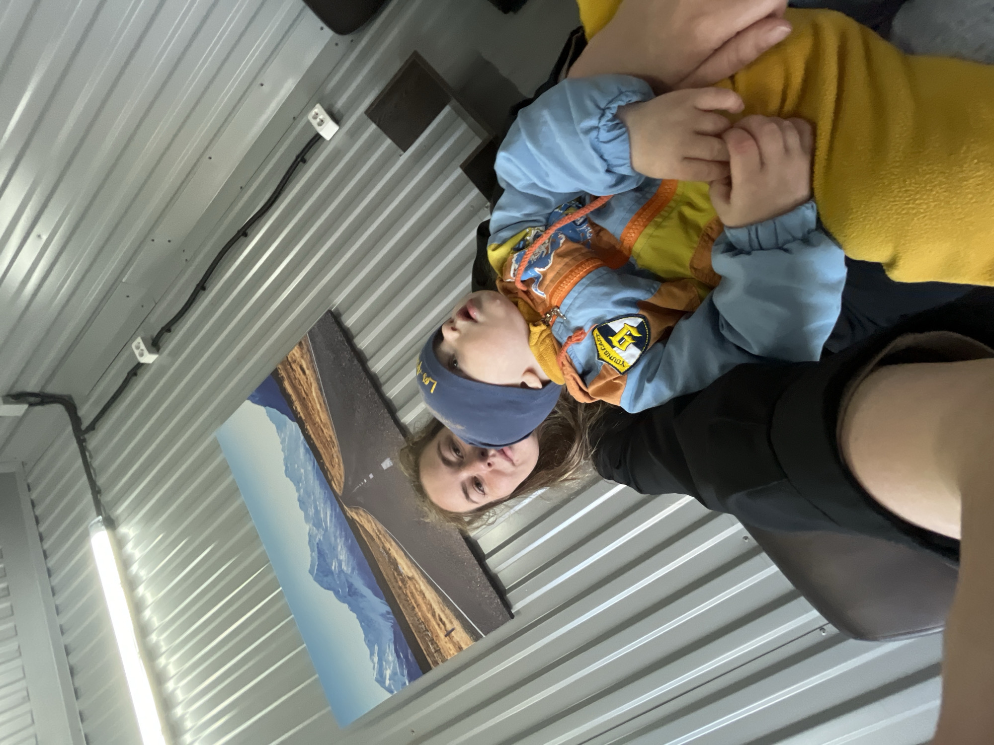 The image shows a person sitting in a room with corrugated metal walls. The person is holding a child wearing a blue hat and a blue and yellow jacket. There is a poster of a road and mountains on the wall. The photo appears to be taken indoors, possibly in a vehicle or a prefabricated structure. It is difficult to determine the exact location as Chaikovsky, Russia from the image alone.