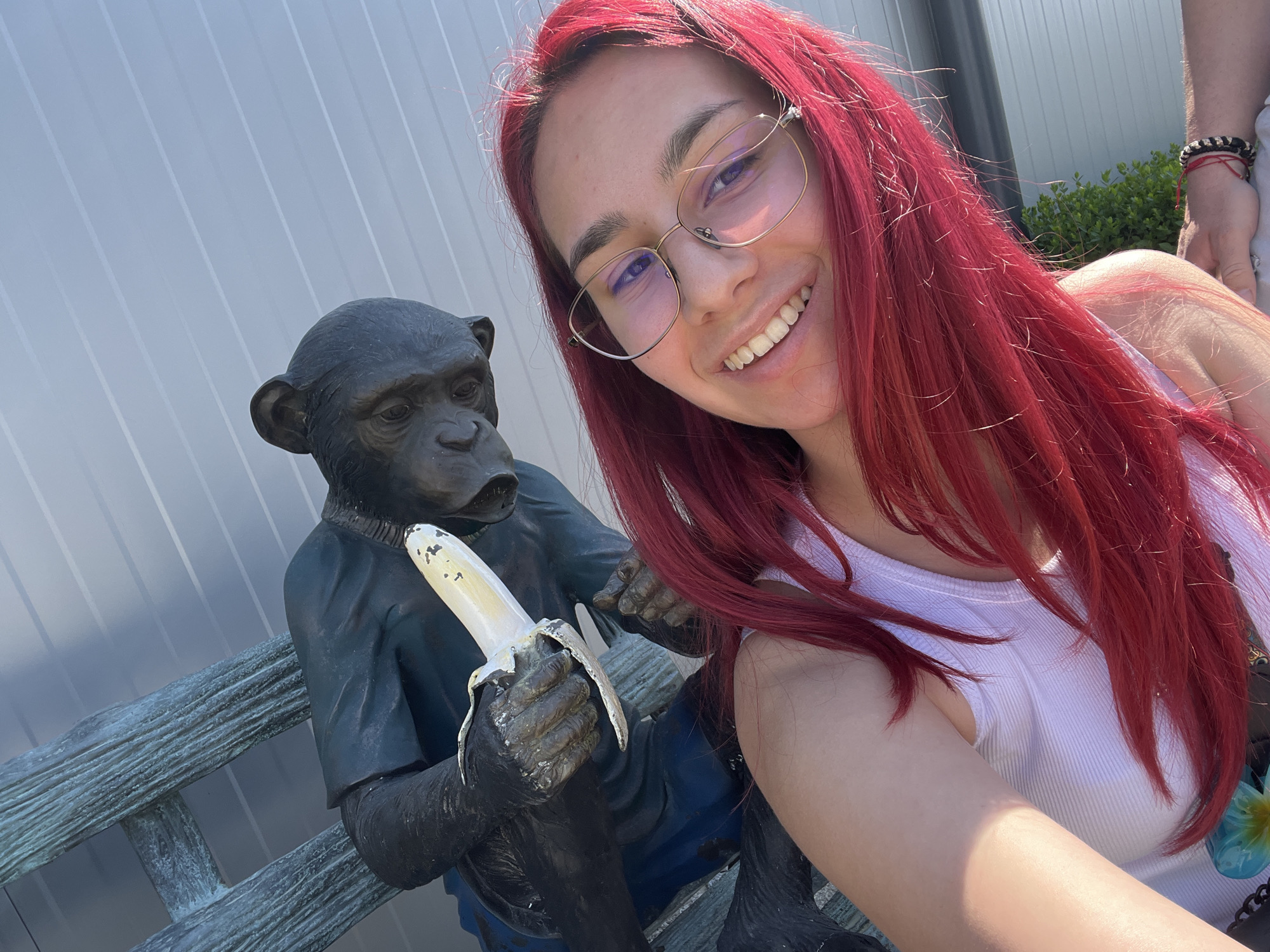 A young woman with red hair and glasses smiles while taking a selfie next to a bronze monkey statue sitting on a bench, holding a peeled banana. The statue is wearing a dark shirt and appears to be peeling the banana. The bench has a weathered, wooden texture. The background consists of a gray wall with vertical lines and a glimpse of greenery and a person's arm on the right side of the frame. The photo was taken in Asenovgrad, Bulgaria.