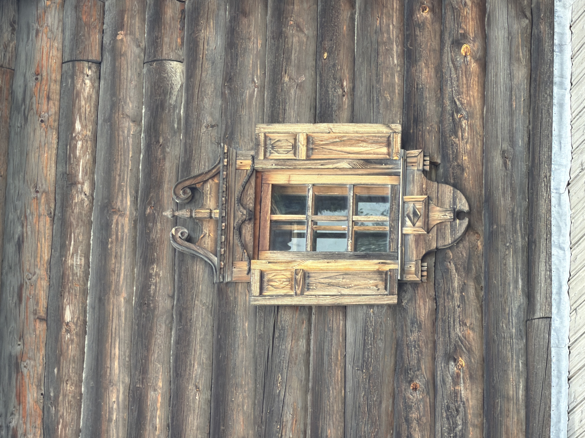 The image shows a close-up view of a rustic, wooden window set into a log wall. The window is adorned with intricate, carved wooden trim, and has shutters that are partially open. The log wall is made of horizontal, weathered logs, showing signs of age and natural wear. The overall impression is one of a traditional, possibly historic, structure. The provided context indicates this scene is located in the city of Imeni Sverdlova, Russia.
