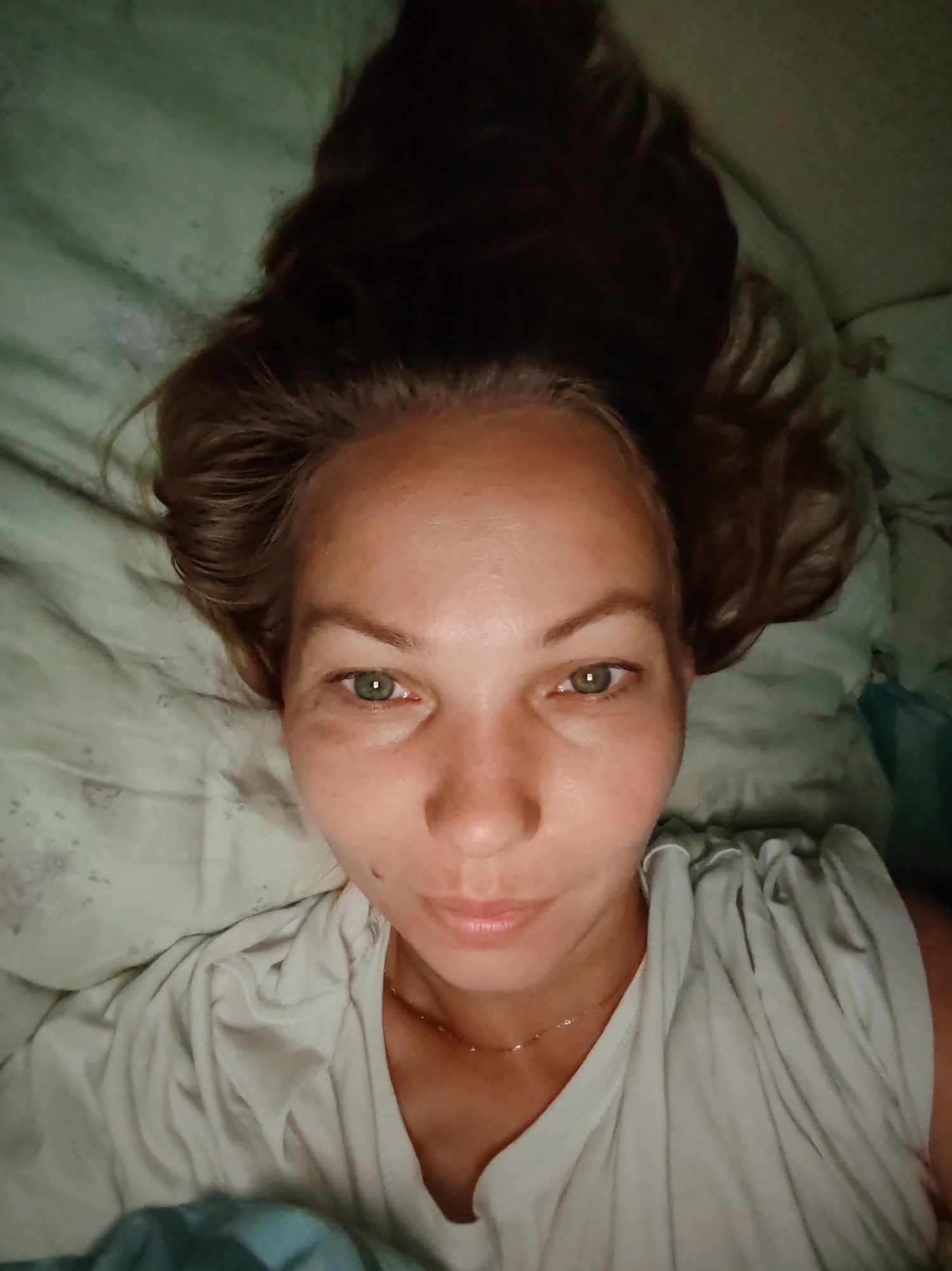 A woman is lying on a bed with her head turned to the side. She has long brown hair that is spread out around her head. She is wearing a white t-shirt and a thin gold necklace. Her eyes are open and she is looking at the camera. The background is a light green sheet. The image appears to have been taken indoors, possibly in a bedroom. There is no indication of the location being in the city of Чухлэм, Russia.