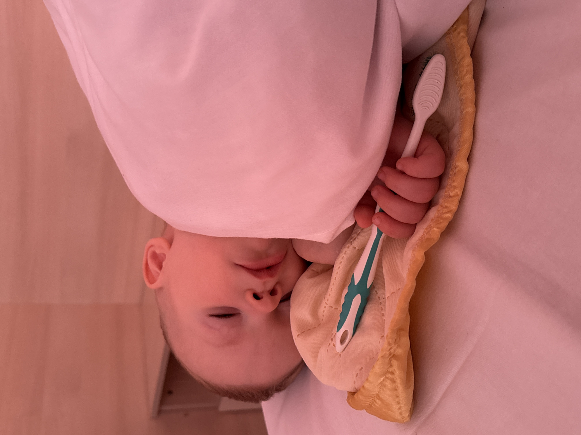 A baby is asleep, wrapped in a pink blanket. The baby's hand is holding a white and teal toothbrush. The baby is lying on a yellow blanket, which is on a white surface. The background is a pink wall and a light-colored shelf. The image appears to have been taken in a bedroom.