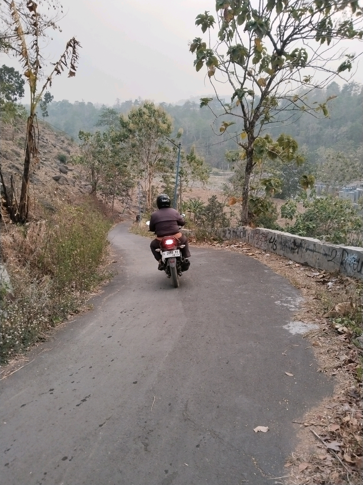 The image depicts a person riding a motorcycle on a paved road that curves to the left, surrounded by dry vegetation and trees. The road has some dark stains on its surface. On the right side of the road, there is a concrete barrier with graffiti. In the background, there is a hazy landscape of hills and trees. The sky is overcast. The license plate of the motorcycle reads "C 3653 YZ". The person is wearing a dark helmet and dark clothing. The location appears to be a rural or semi-rural area, potentially in Sangkanjaya, Indonesia, given the context provided.
