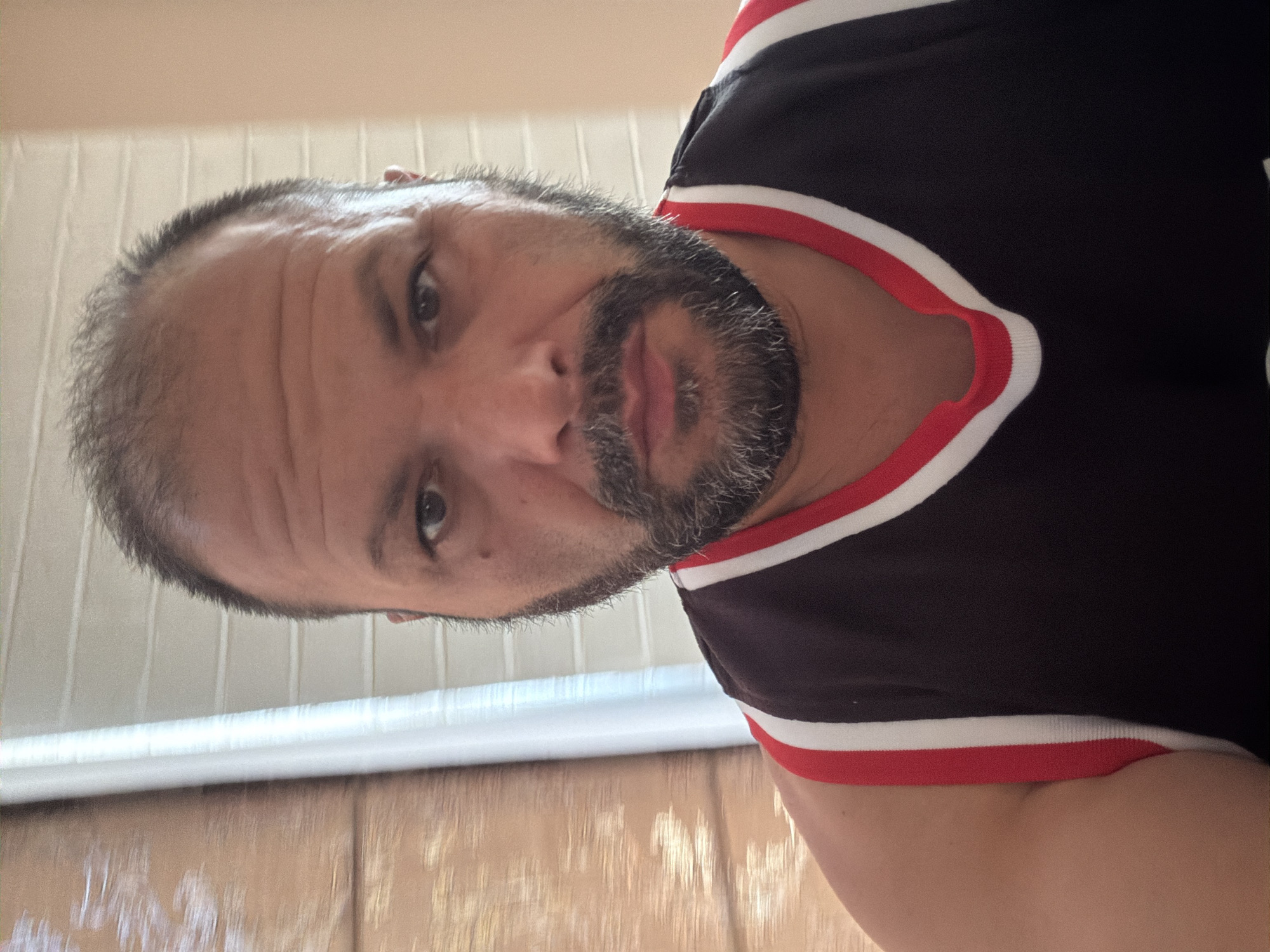 A medium shot photograph shows a man in his 30s or 40s taking a selfie. He has a receding hairline with short, dark hair and a salt-and-pepper beard. He is wearing a sleeveless black jersey with red and white trim around the neckline. The man's left arm is visible on the left side of the frame. Behind him, on the left, is a wooden blind or shutter with vertical slats. To the right of that is a white vertical pole, possibly part of a window frame. The wall behind the man on the right is painted a light peach color and appears to have horizontal paneling. The lighting suggests an indoor environment, possibly a home, with natural light coming from the left. The man's expression is neutral. The image does not provide enough information to determine a specific location within the city of Burgas, Bulgaria.