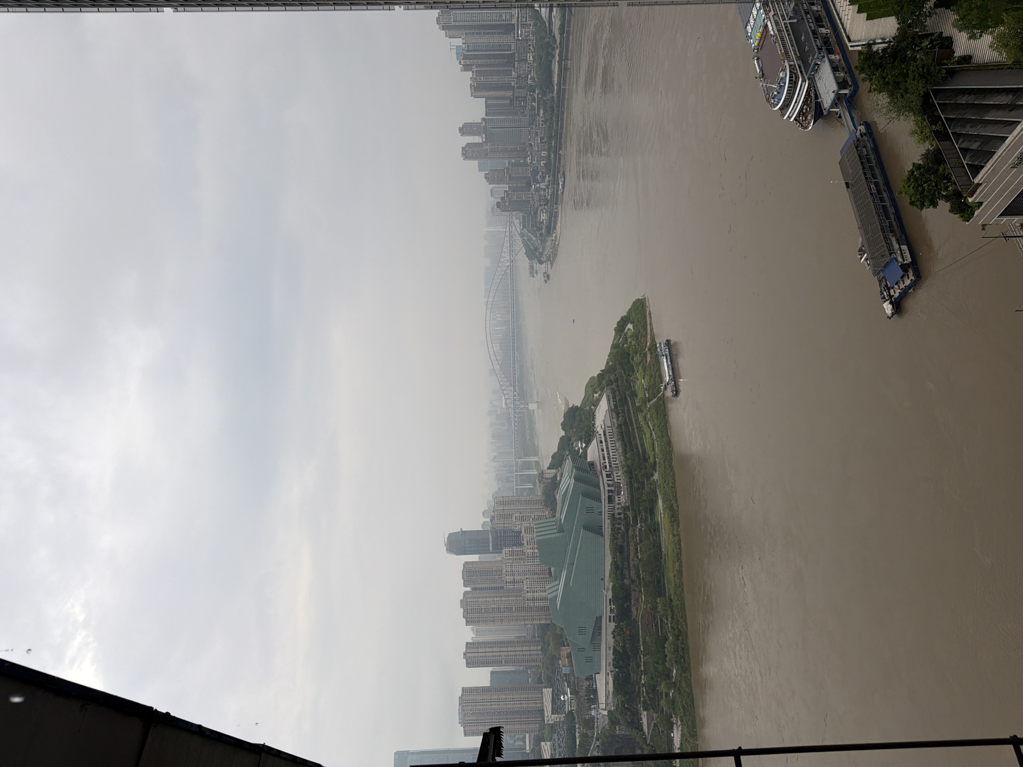 The image shows a panoramic view of the confluence of the Jialing River and the Yangtze River in Yuzhong District, China, under a cloudy sky. A large, modern concert hall with a distinctive angular green roof is situated on a promontory on the left side of the frame, bordered by trees and landscaping. Across the muddy brown river, a dense cityscape of tall buildings rises, with several bridges spanning the water in the distance. Two ferries or tour boats are docked on the right side of the river in the foreground. The overall scene depicts an urban riverfront with significant infrastructure and architectural development.
