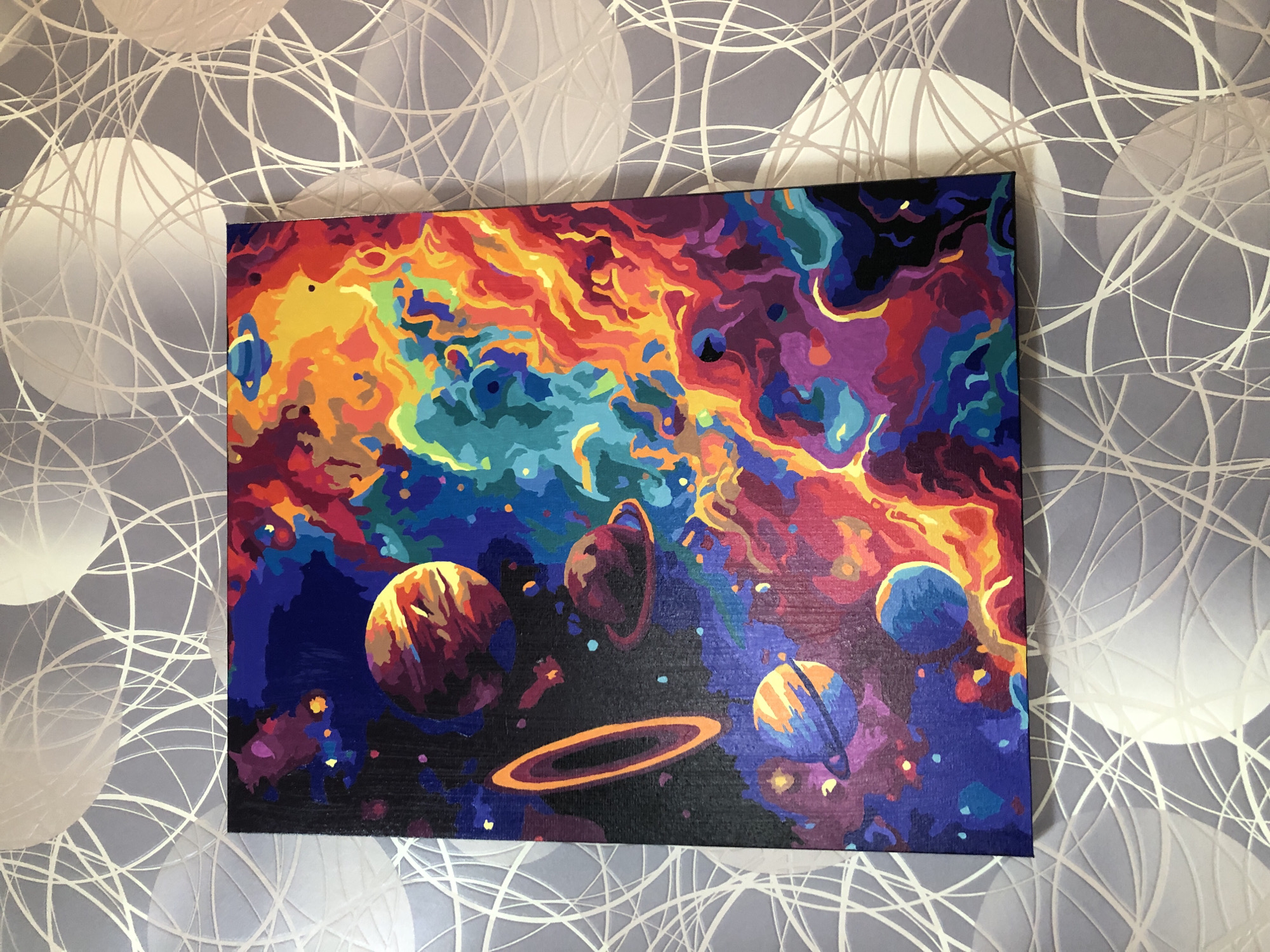 The image displays a painting of a vibrant, colorful nebula with planets. The painting is positioned on a wall with a patterned wallpaper that features white circular shapes and thin white lines. The painting itself is a vivid depiction of space, with swirling masses of red, orange, yellow, blue, and purple. Several planets are visible, some with rings. One prominent planet in the upper left has orange and yellow stripes, while another below it is blue with an orange ring. There is also a thin, orange oval shape that could represent a ring or a celestial object. The background of the painting is a dark, starry expanse. The wallpaper behind the painting has a textured appearance and a greyish-blue hue. The painting is rectangular and oriented vertically. No people or specific locations in Safonovo, Russia are visible in the image.