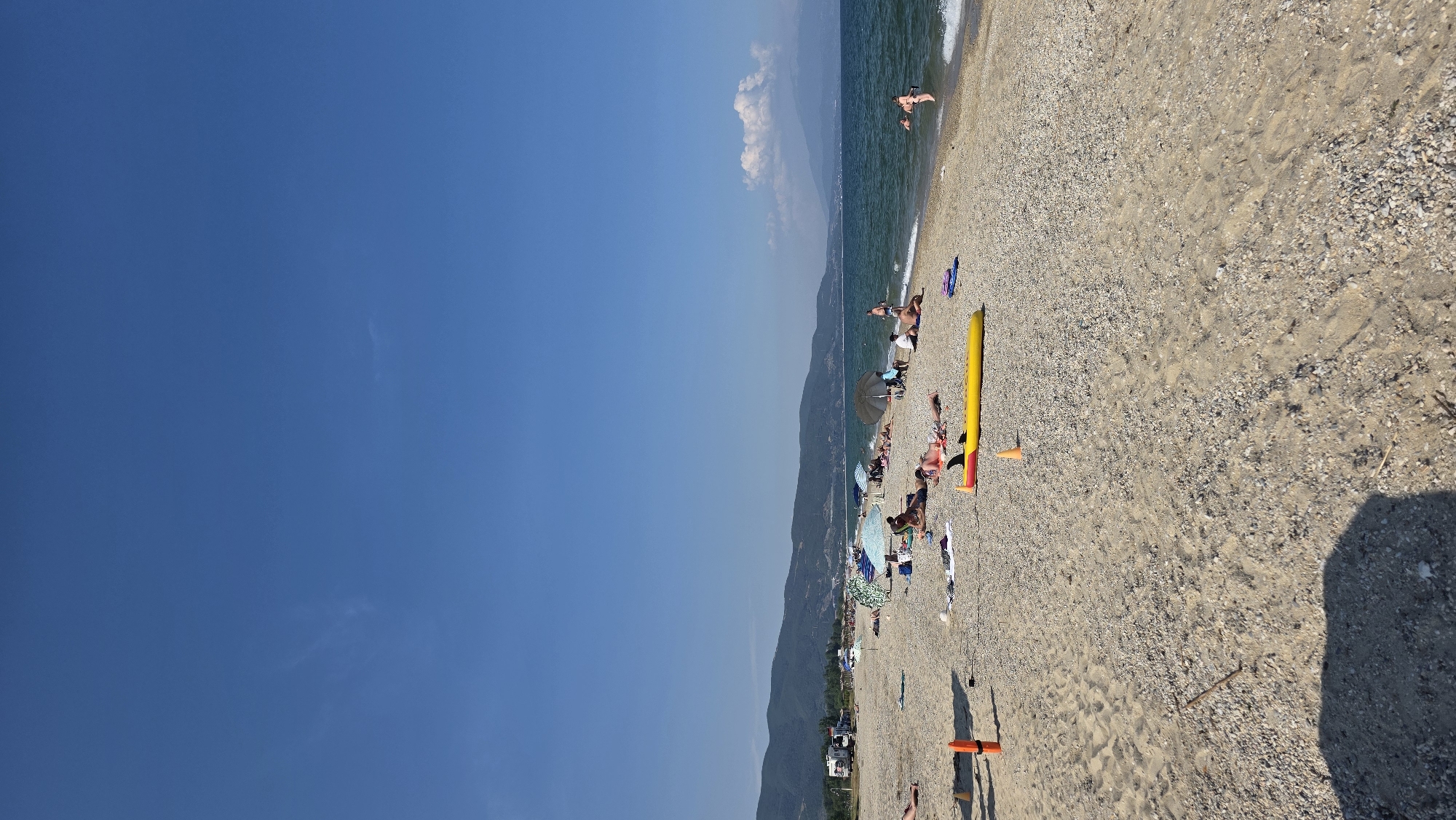 The image captures a sunny day at a pebble and sand beach in Ασπροβάλτα, Greece. The sky is bright blue with faint high-altitude clouds. A range of green, tree-covered mountains dominates the background, with distant peaks and a large white cumulus cloud visible on the far right. The sea is a dark blue-green, exhibiting small waves breaking gently at the shore.
Approximately 20-30 individuals are present on the beach and in the water. Many are seen sunbathing on towels or directly on the sand, while others are seated under various beach umbrellas (blue, green, and dark grey). Two individuals are partially submerged in the water on the right side, one appearing to enter and another further out.
Key objects on the beach include a prominent yellow inflatable object, possibly a paddleboard or raft, with black fins, positioned in the mid-ground. Two small orange traffic cones are also visible on the sand. A blue pair of sandals rests near the yellow inflatable. In the far left background, a white recreational vehicle (RV) and other vehicles are parked near the base of the mountains. A large, dark shadow from an unseen object extends into the lower left foreground.