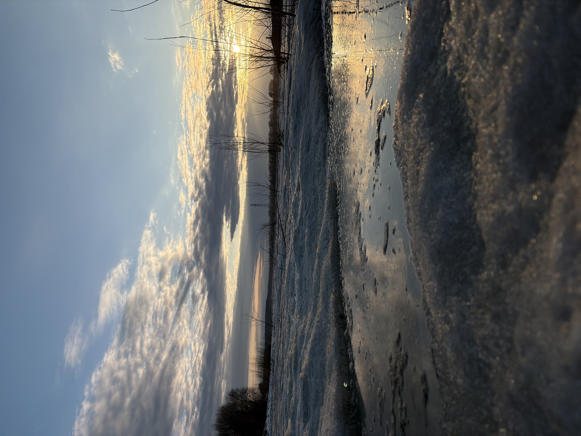 The image shows a winter scene, possibly taken at ground level, featuring a partially frozen body of water reflecting the sky and sunset. The sky is filled with clouds, some illuminated by the setting sun, creating a warm, golden light. Bare trees and bushes line the horizon, silhouetted against the bright sky. The foreground consists of ice and snow with textured surfaces, indicating melting and refreezing. Based on the prompt, the location is Chaikovsky, Russia. There are no people visible in the frame. The primary subjects are the winter landscape, the frozen water, and the sunset sky. The main action is the sunset casting light onto the environment.