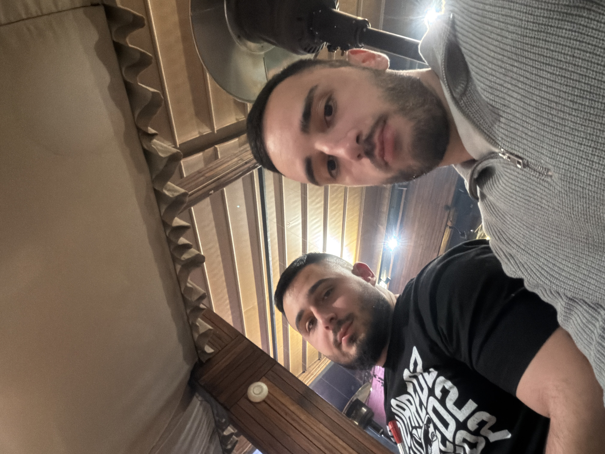Two men are visible in the image, likely a selfie. The man at the top of the frame is wearing a gray zip-up sweater. The man at the bottom is wearing a black t-shirt with a white design. They both have dark hair and beards. The background appears to be an indoor space with textured ceiling panels and wooden beams. There is a light fixture above the man in the gray sweater. Based on the architecture, the location might be a restaurant or bar in Plovdiv, Bulgaria. There are no discernible actions or interactions.
