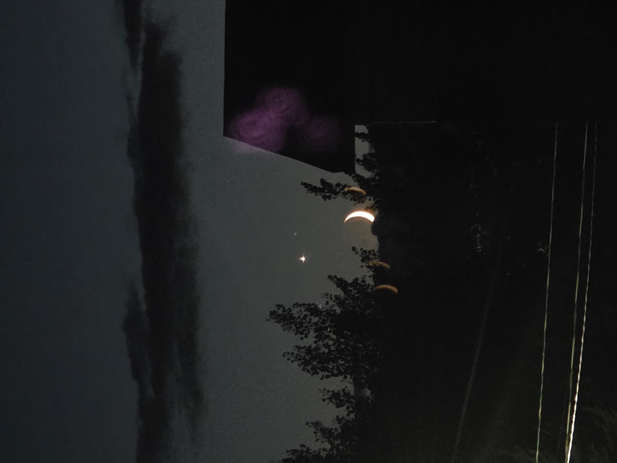 This is a nighttime photograph taken in the city of Bryansk, Russia. The scene is dark, with a sliver of a crescent moon visible in the sky. A bright star or planet is also visible near the moon. The foreground is dominated by the silhouette of trees and a portion of a building. The building appears to have a dark roof and some faint, diffused purple light on its side. There are also power lines visible in the lower part of the image, which are illuminated by a light source, possibly streetlights, casting a greenish-white glow. The overall impression is of a twilight or early night sky with celestial objects and urban infrastructure elements.