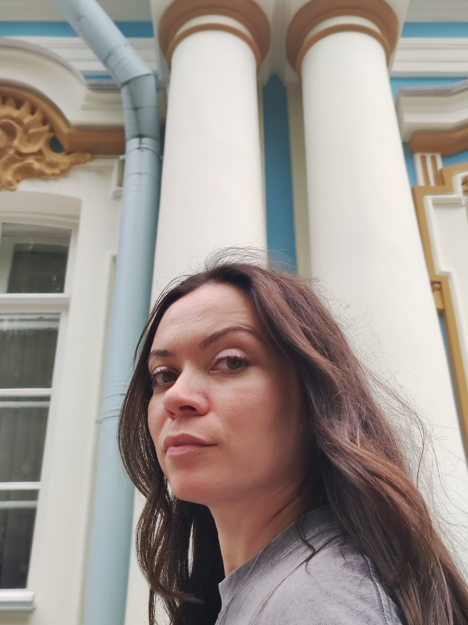 An outdoor, low-angle medium shot captures a woman with long, dark brown hair looking directly at the viewer with a serious expression. She is positioned in the foreground, her face taking up a significant portion of the frame. Her skin appears fair, and she has dark eyebrows and lashes. She wears a gray, collared shirt. In the background, a light-colored building with decorative elements and large white columns with gold trim is visible. A light blue downspout runs vertically along the building. The architecture suggests the location is likely in Pushkin, Russia, possibly near one of its palaces or grand estates.