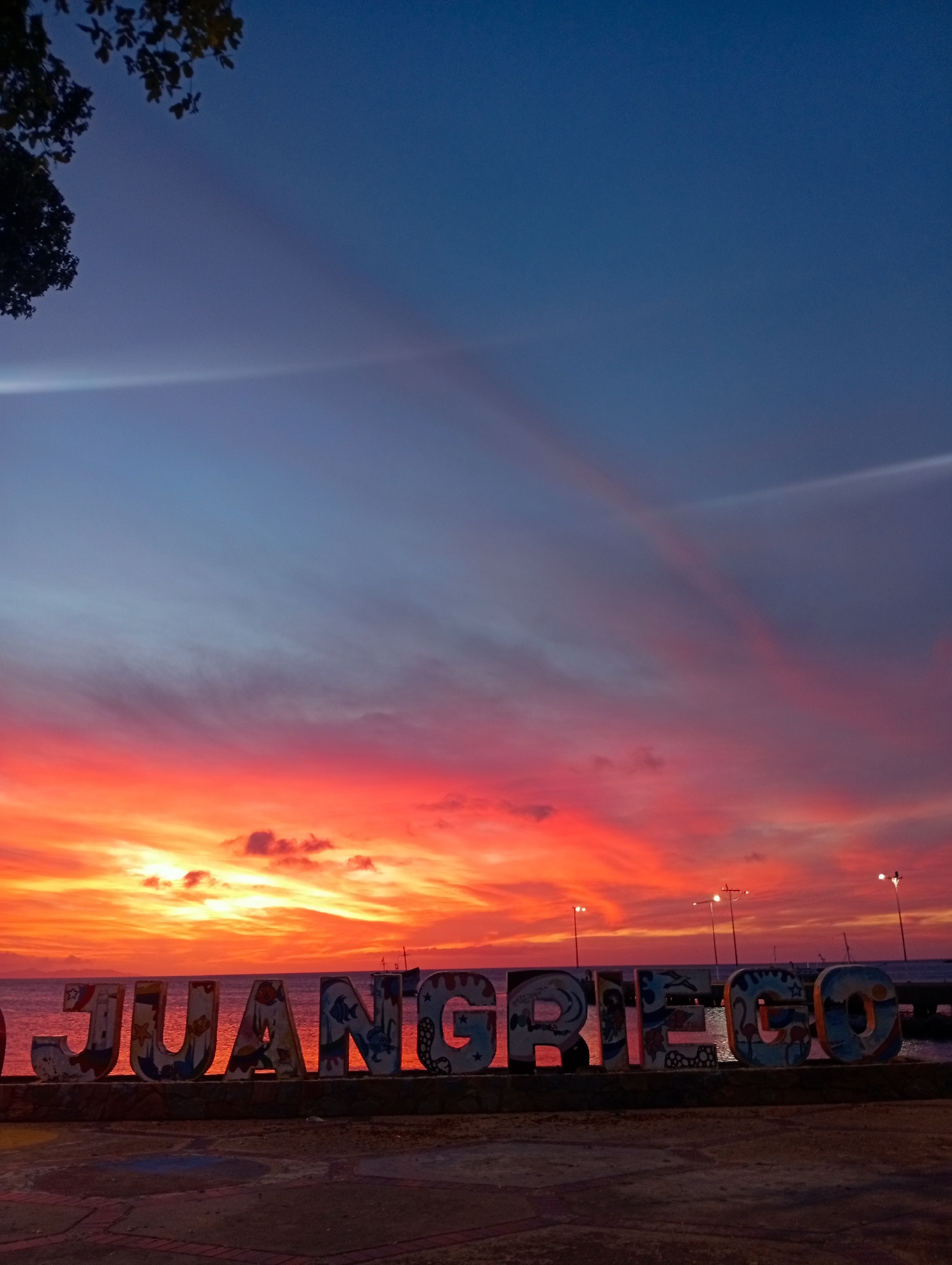 The image depicts a twilight scene in Juan Griego, Venezuela, specifically at a waterfront promenade. A large, multi-colored sign spelling "JUAN GRIEGO" stands prominently in the foreground, constructed from individual, decorated block letters. The letters are painted with various designs, including marine life and symbols. The sign rests on a dark, textured base.
Behind the sign, a body of water, likely the ocean or bay, is visible, reflecting the ambient light. The horizon is dominated by an intense sunset, displaying a vibrant gradient of deep orange, red, and yellow hues transitioning into purple and then dark blue in the upper sky. Scattered clouds are present, some illuminated by the setting sun, while others appear as darker silhouettes. Two distinct light streaks, possibly contrails or atmospheric phenomena, are visible diagonally in the upper sky.
On the far side of the water, several streetlights on poles are illuminated, casting bright, localized light. To the right of the "JUAN GRIEGO" sign, a dark silhouette of what appears to be a moored vessel or ship is discernible against the brightly colored horizon. In the upper left corner, dark foliage from a tree is visible, providing a natural frame. The immediate foreground shows a paved or tiled ground surface, partially illuminated by the residual light. No individuals or active interactions are observed in the scene.