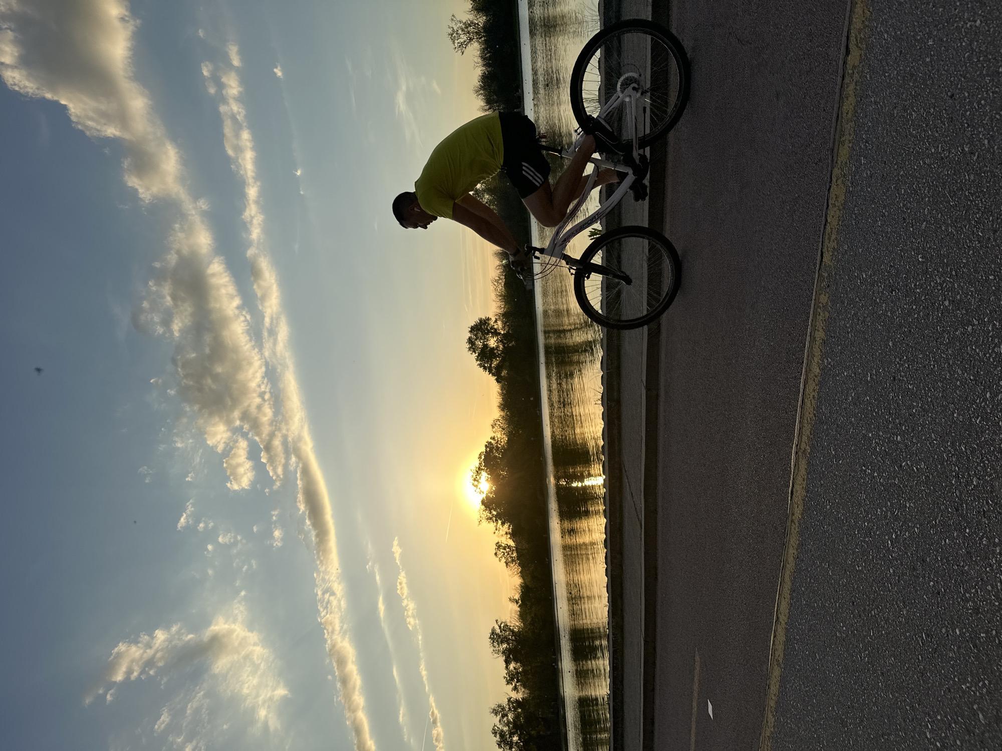A photograph, oriented vertically, shows a man performing a bicycle trick on a paved road near a body of water. The man is positioned in the center of the frame and is wearing a green shirt and dark shorts. He is bent over the handlebars of his white bicycle, with the front wheel elevated slightly off the ground. The bicycle appears to be a mountain bike style, with knobby tires.
The scene takes place at dusk or dawn, with the sun setting or rising just above the trees in the background, casting a warm glow over the water. The water appears calm and reflective. Trees line the opposite bank of the body of water. The sky above is filled with streaks of clouds.
The paved road is on the right side of the image and has a yellow line marking its edge.
This photograph was most likely taken near the Rowing Canal in Plovdiv, Bulgaria.
