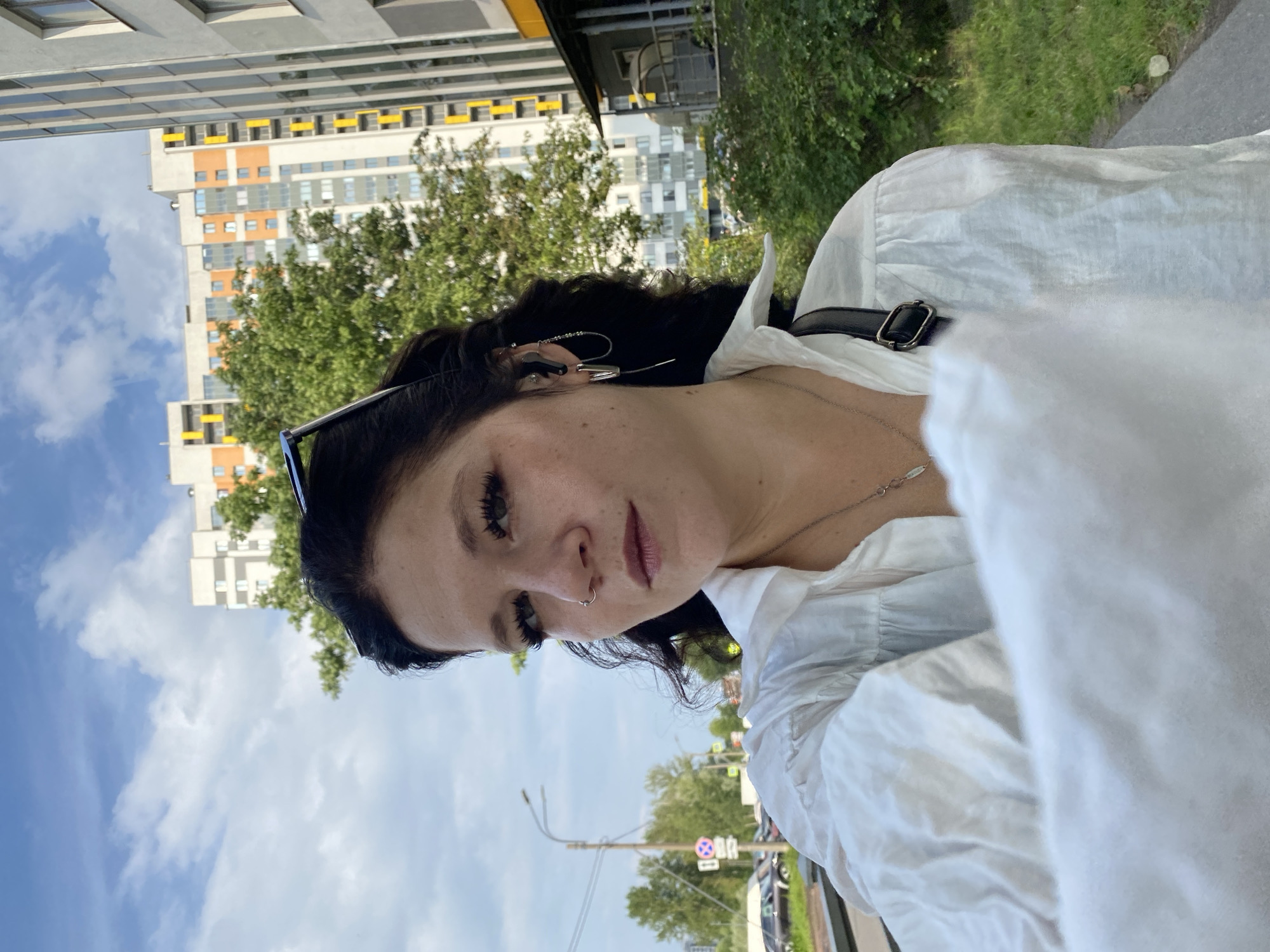 A woman with dark hair is standing outside, looking at the camera. She is wearing a white blouse and has on a nose ring and earrings. A black bag strap is visible over her shoulder. In the background, there are apartment buildings, trees, and a cloudy blue sky. The scene appears to be in an urban environment.