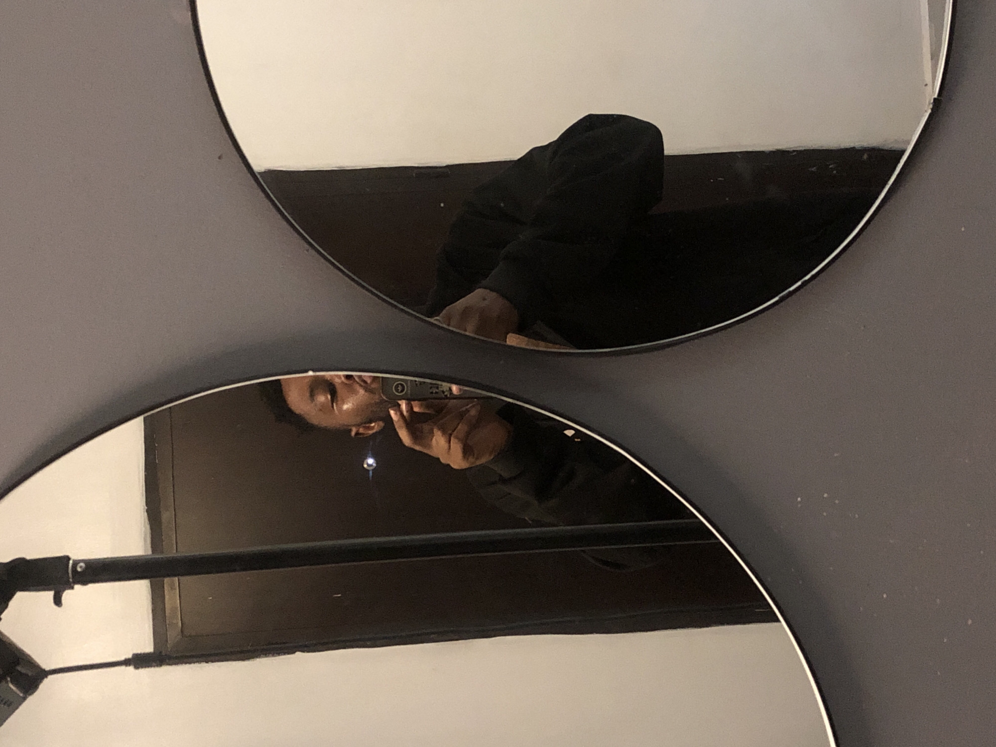 The image captures a person's reflection in two circular mirrors mounted on a dark gray wall. The mirrors are arranged vertically, with the top one showing a partial view of a reflection and the bottom one presenting a clearer, inverted reflection of a person.
In the lower mirror, a person with dark skin is reflected, appearing upside down. They are wearing a dark-colored garment, possibly a hoodie. Their face is visible, and they are holding a smartphone in their right hand, seemingly in the act of taking a photograph, likely a selfie or a shot of the mirrors. A small, bright light point, possibly from the phone's flash or another light source, is visible near their head in the reflection. A dark, horizontal bar-like object is also reflected across the lower part of this mirror.
The upper mirror shows a more obscured reflection, primarily of the person's arm and torso, also clad in a dark garment. The orientation of this reflection suggests a different angle or perspective from the person's body relative to the upper mirror.
The reflections within both mirrors are predominantly dark, hinting at a dimly lit environment or the dark attire of the subject. The wall surrounding the mirrors is a solid, dark gray color, with no visible text or distinctive markings. The scene depicts an indoor setting, and the primary activity is the taking of a photograph using mirror reflections.