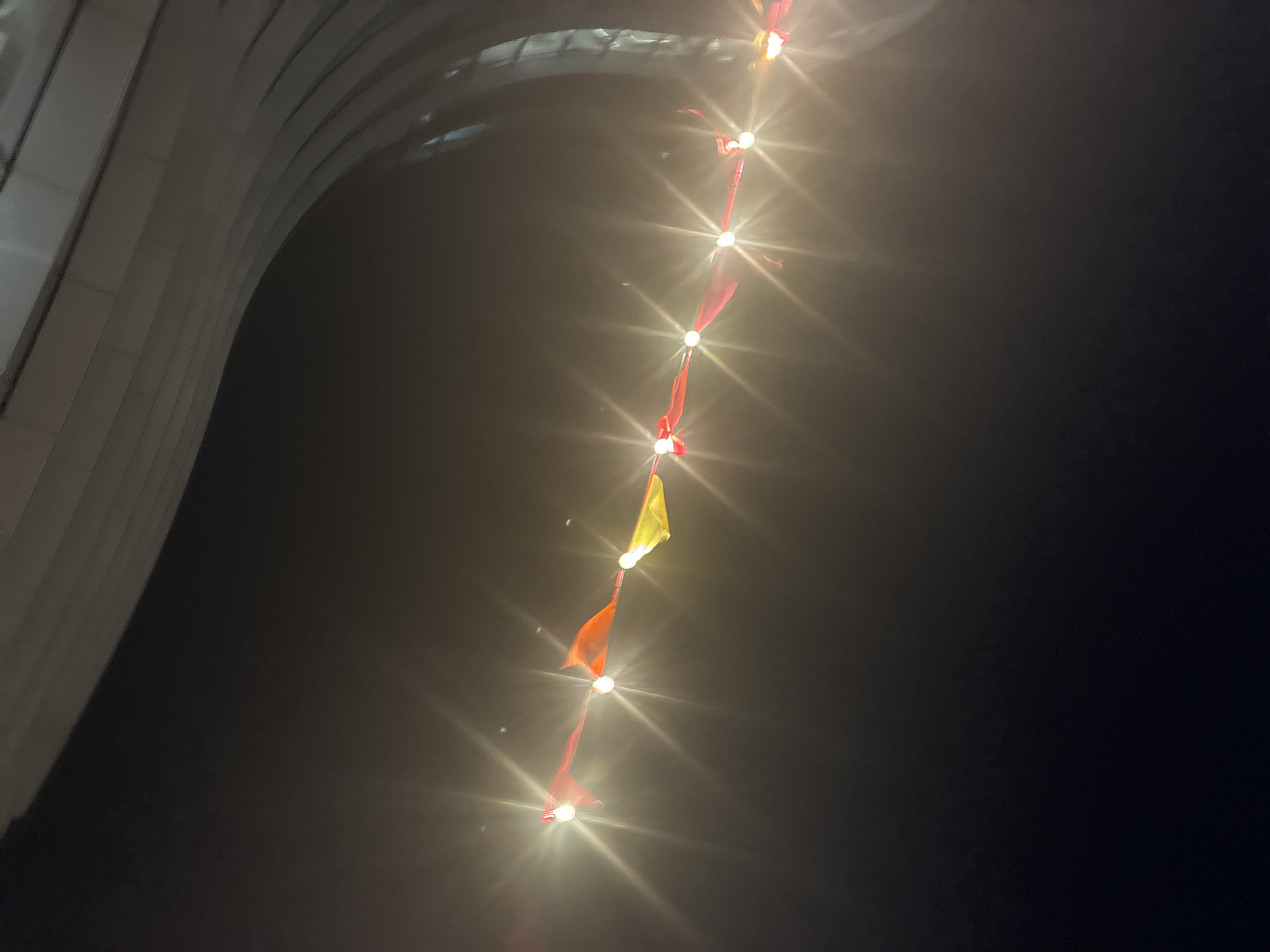 The image shows a string of lights hanging near a structure in London, United Kingdom. The structure appears to be part of a building or monument, its architectural details visible on the left side of the frame. The lights are strung vertically, creating a line of illumination against a dark background, which likely represents the night sky. Attached to each light is a colored flag, primarily red, with one flag being yellow. The image is taken from a low angle, looking upwards towards the lights and the structure. The overall scene is dimly lit, with the lights providing the primary source of illumination. The atmosphere appears to be foggy or misty, contributing to the diffused glow around the lights.
