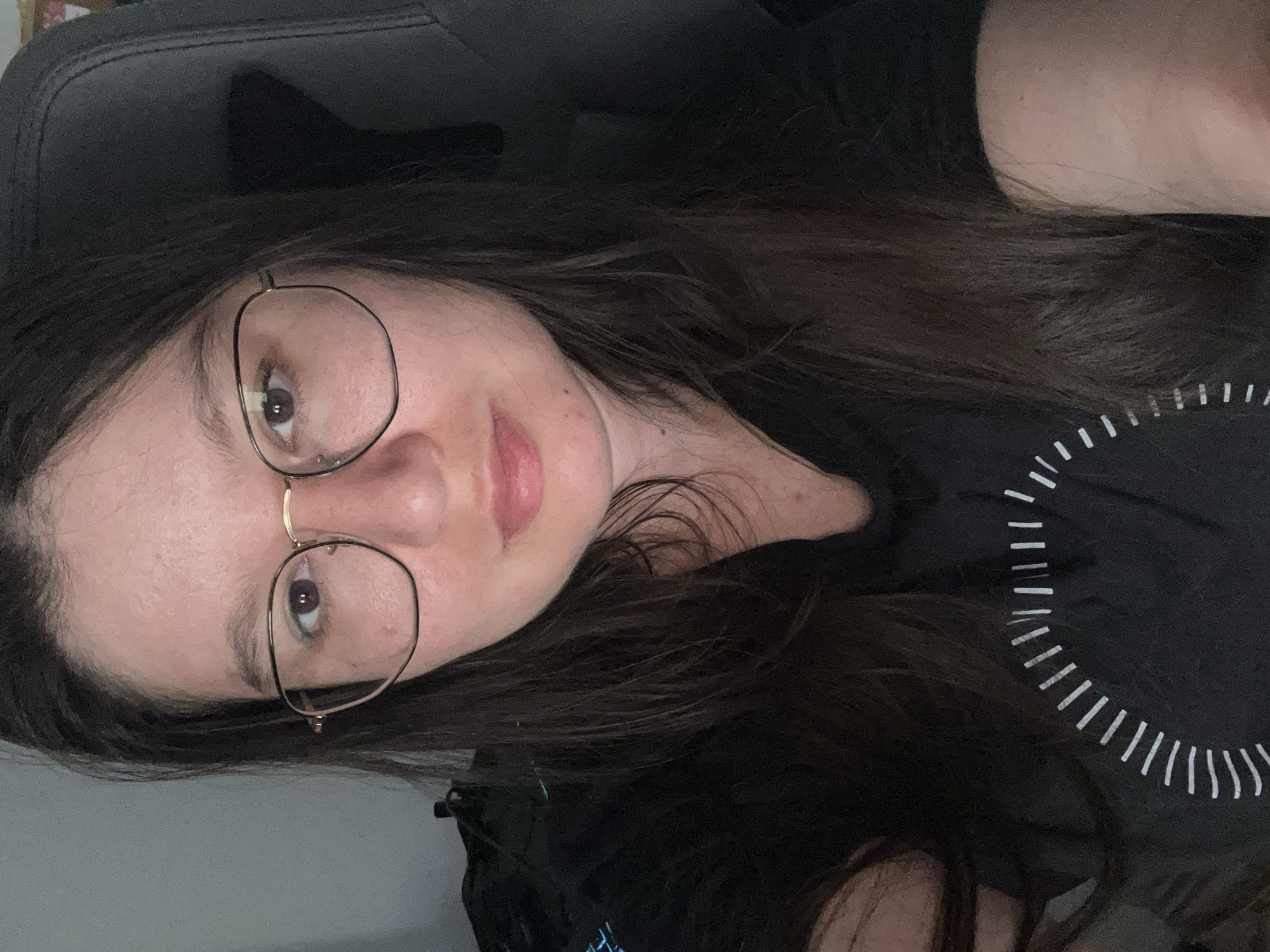 A person with long brown hair and eyeglasses is facing the camera. The person is wearing a black shirt with a white circular design. The person is seated in a black chair. The location is not discernible from the given image.