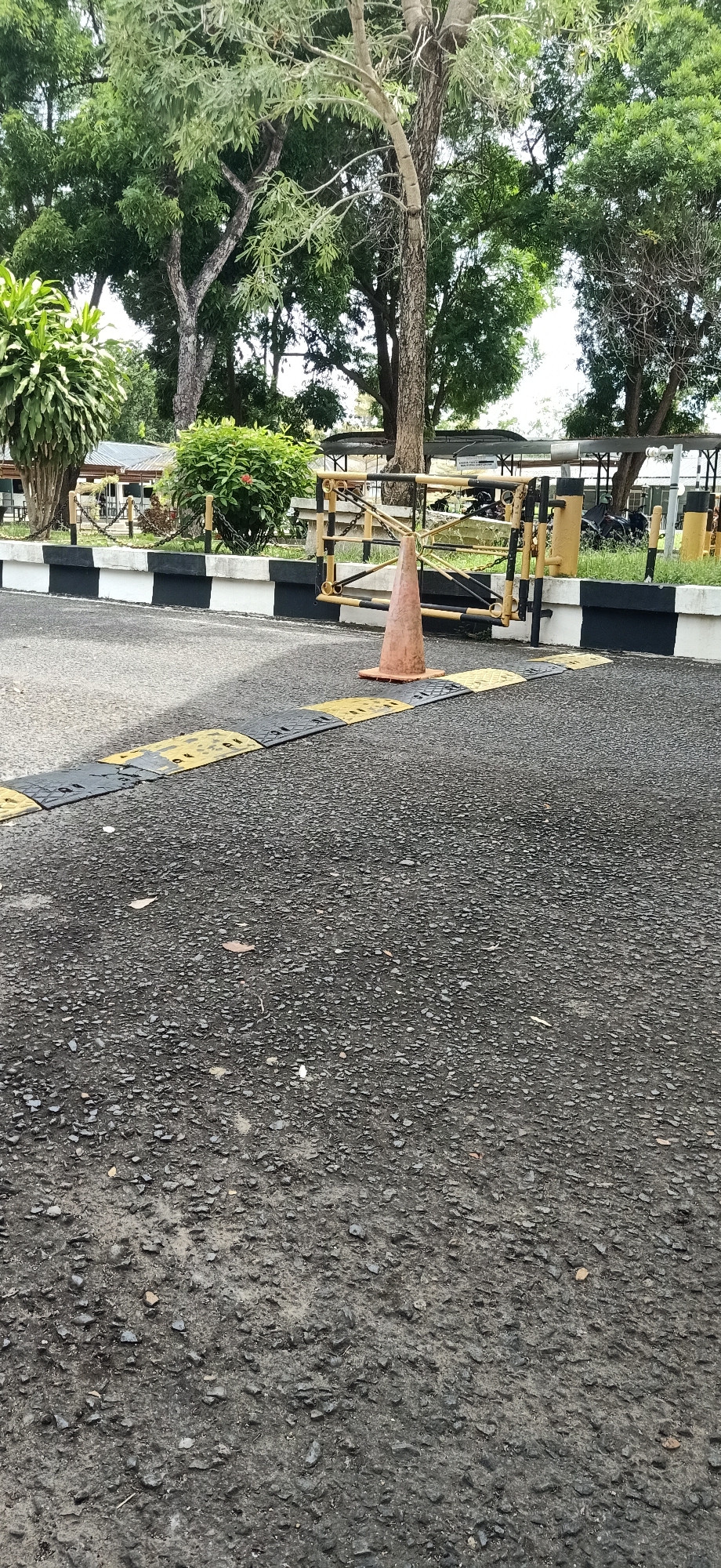 The image depicts a road with speed bumps in Tambontunong, Indonesia. A traffic cone is placed on the road near a yellow and black striped barrier. In the background, there are trees, vegetation, and some buildings. The scene appears to be a street or an entrance to a facility. There are no people visible in the image.