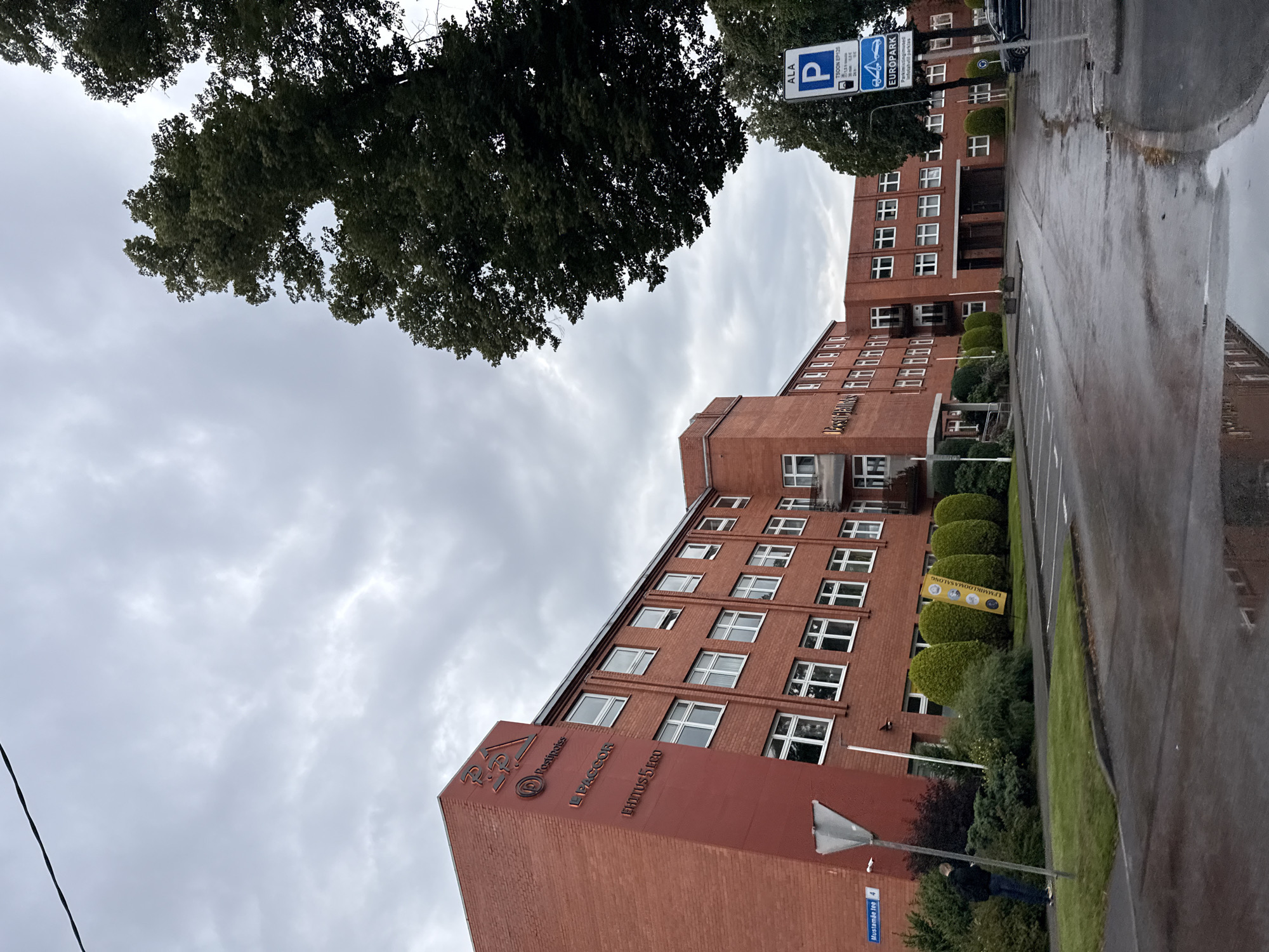 The image captures an exterior view of a brick building in Tallinn, Estonia, on a cloudy day. The building, with multiple windows and a dark roofline, appears to be an office or commercial space. Several signs are visible on the building's facade, including "P.R. Postipois," "LAPPCOR," and "EHITUS 5 tee." A street sign indicates "Mustamäe tee 4." In front of the building, there is a wet parking lot with a few cars and a person with a backpack standing near the sidewalk. Lush green bushes and small trees line the property. To the right, a tall tree overhangs the scene, and a parking sign for "EUROPARK" is also present. The overall impression is of an urban setting with overcast weather.
