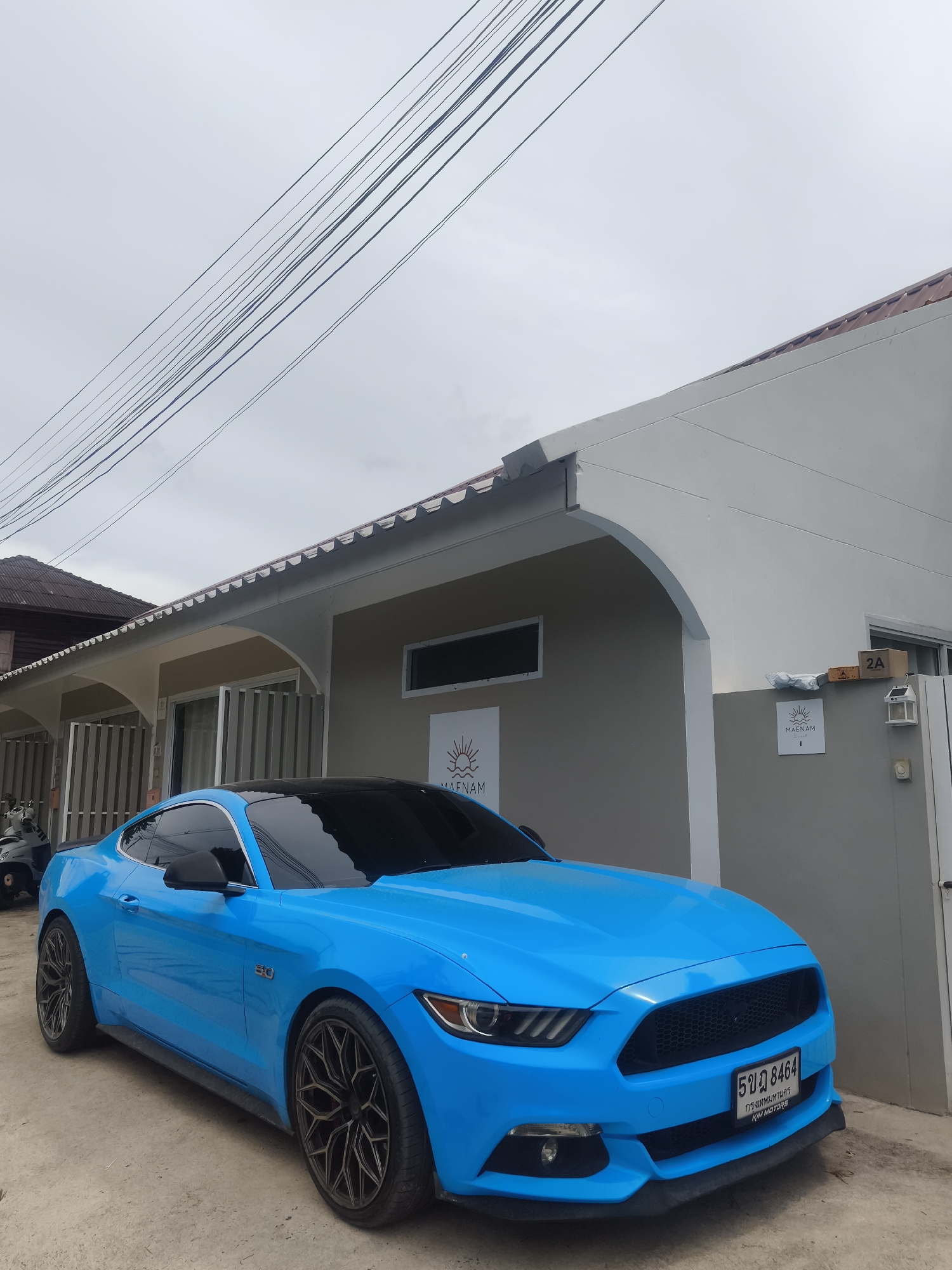 An outdoor, eye-level, medium shot captures a bright blue Ford Mustang parked in front of a neutral-toned building under an overcast sky. The car, a coupe with a black roof and tinted windows, faces slightly to the left, showcasing its front grille, headlights, and license plate. The license plate, with Thai characters and numbers, is visible on the front bumper. To the right of the car, a white building features an archway entrance and a sign with "MAENAM" and a sun logo. Above the building, power lines crisscross the sky. The ground in front of the car is a dirt and gravel surface. The scene appears to be on a street in an urban environment.