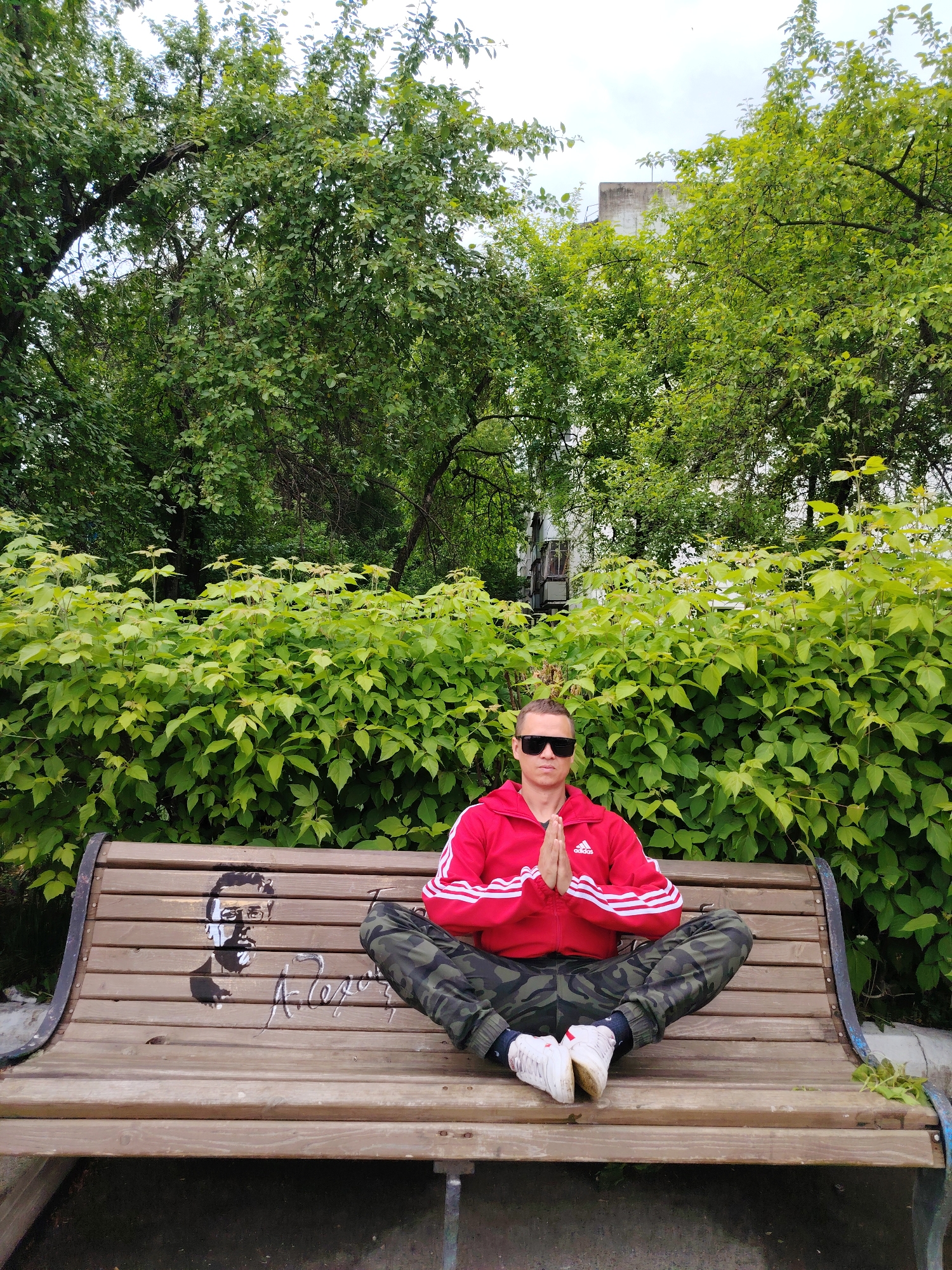 A man sits in the lotus position on a park bench in what appears to be a park in Chaikovsky, Russia. The man wears sunglasses, a red Adidas jacket with white stripes, camouflage pants, and white sneakers. He has his hands in prayer position. The wooden bench has a painted portrait and signature of a man in black paint. Lush green foliage and trees are visible behind the bench. The sky is overcast.