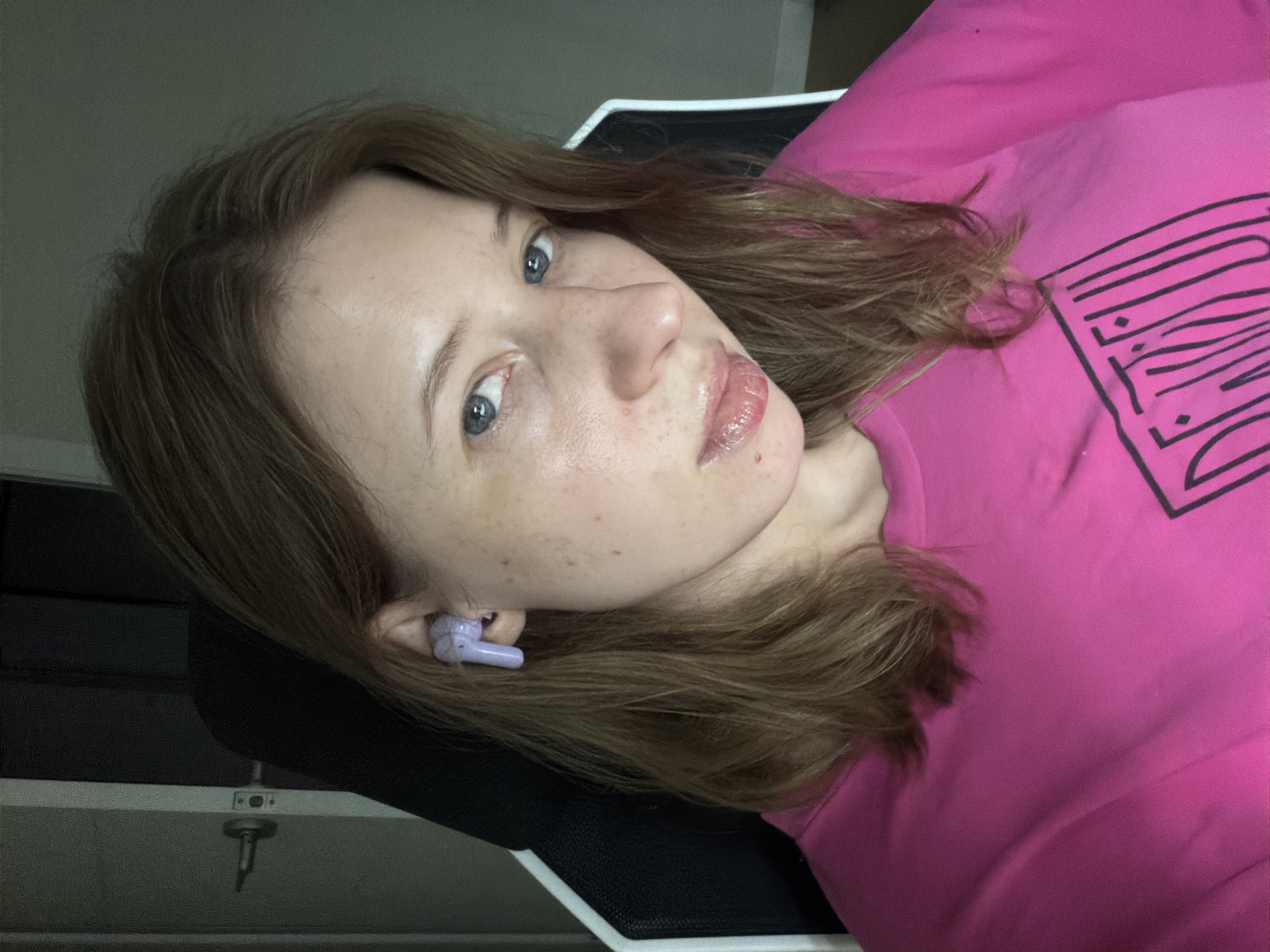 A young woman with shoulder-length brown hair and blue eyes is looking at the camera. She is wearing a bright pink t-shirt with a black graphic design on it. She has a single light purple earbud in her left ear. Her skin has a scattering of freckles. She is seated in what appears to be a gaming chair, with a dark fabric back and white trim. In the background, a door with a silver knob is visible to the left, and the wall behind her is a neutral gray. The lighting in the room is dim. There is no information about the city of Dongducheon-si, South Korea included in this image.