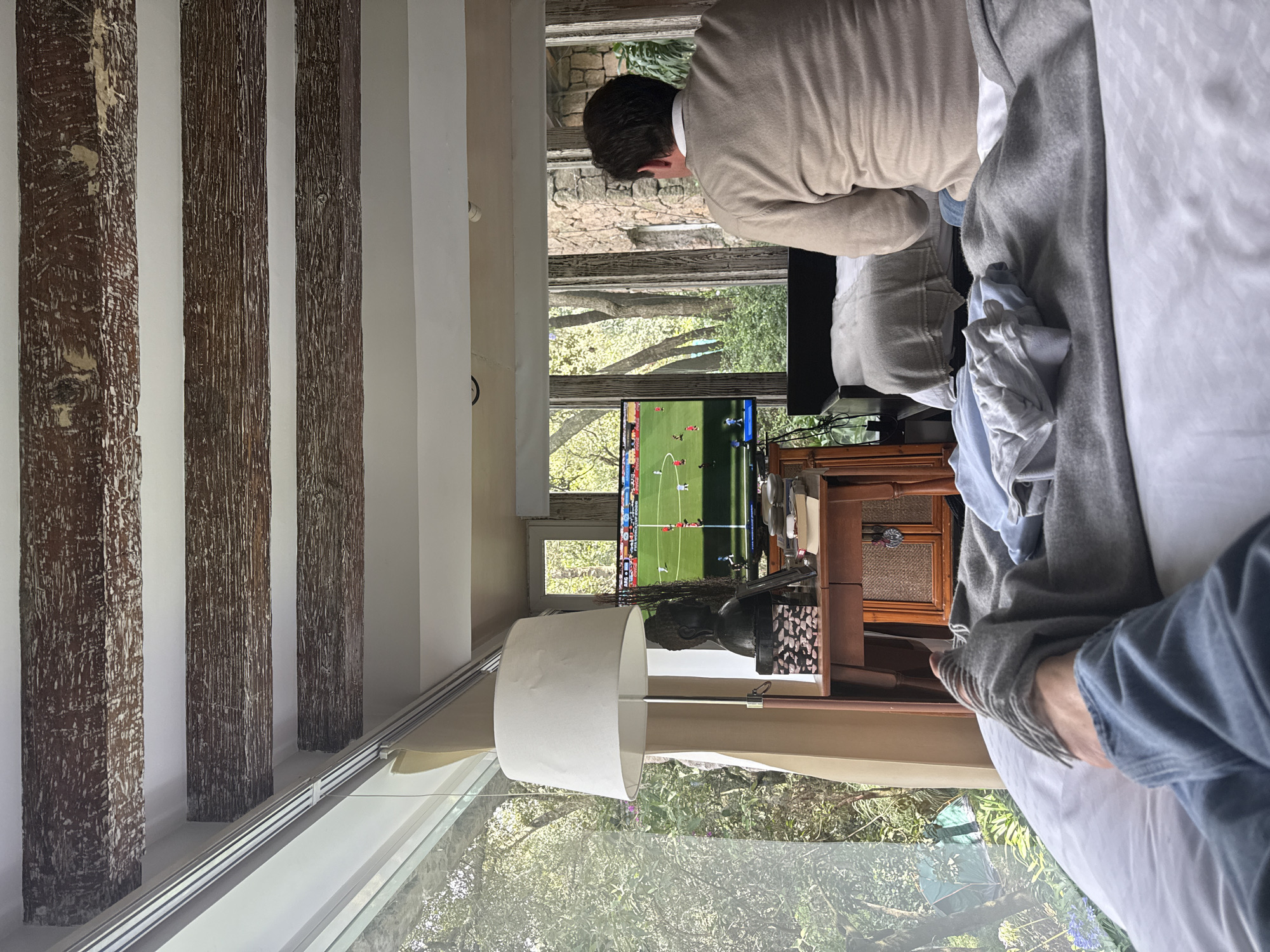 The image depicts an indoor scene within a home, likely in Mexico City, Mexico, with a focus on leisure activities. In the foreground, a person's legs, clad in blue denim jeans and a gray sock with fringed details, are visible resting on a white bed. Adjacent to their legs are neatly folded gray and light blue garments.
The mid-ground is dominated by a television screen displaying a soccer match, identified by "FIFA WORLD CUP 2020" text. A person, from the back, is seated on the edge of a bed, facing the television. They are wearing a beige sweater. Beside the television, a small wooden table holds various objects, including a lamp with a white shade, a dark-colored bust sculpture, a decorative box, a framed photograph, and books.
The background reveals a large window or glass wall offering a view of lush green foliage and trees, suggesting an outdoor environment. The architectural style features exposed wooden beams on the ceiling, adding a rustic element to the interior. The overall impression is a relaxed setting where individuals are enjoying entertainment within a comfortable domestic space.