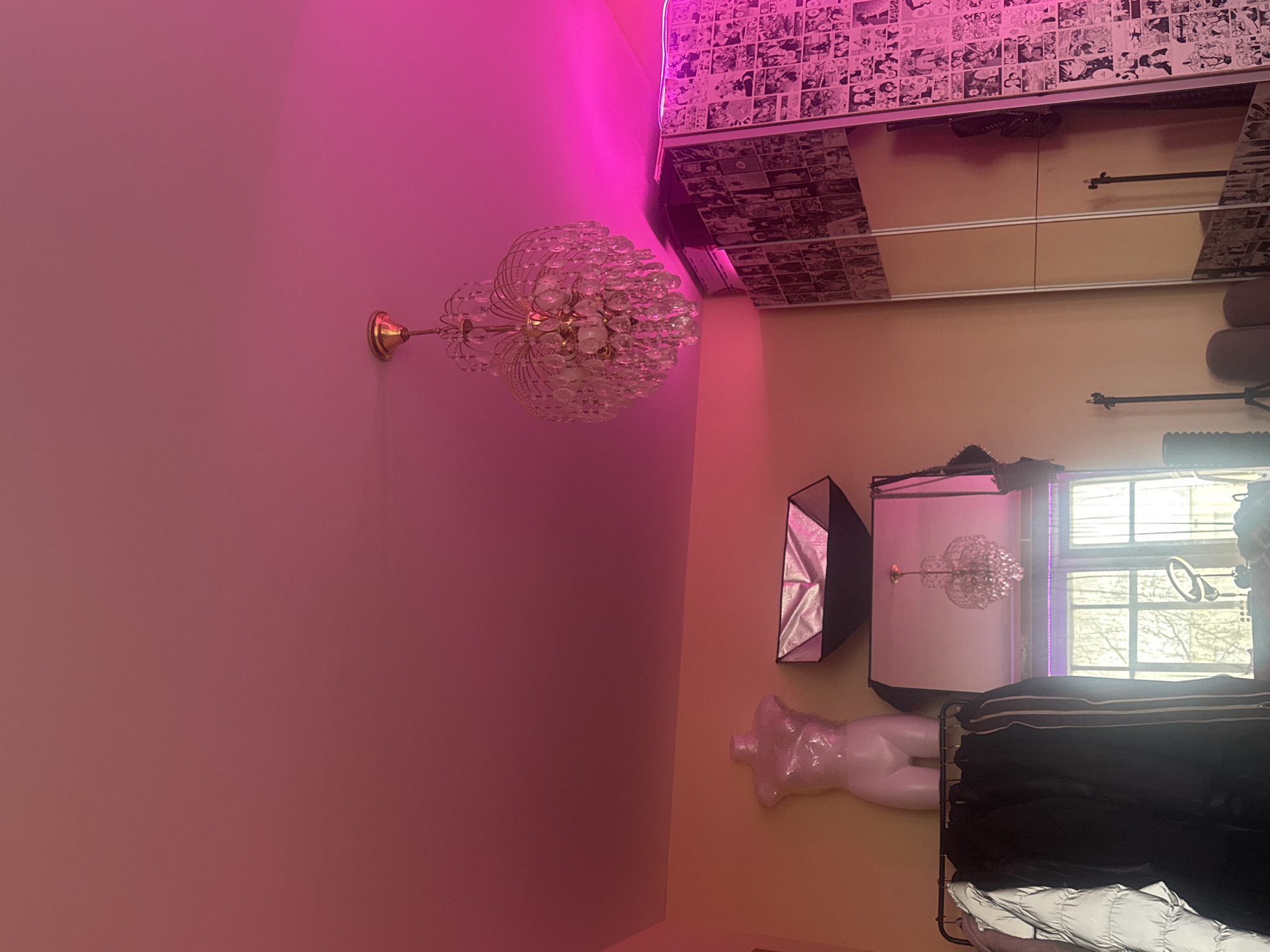 The image depicts an interior scene with elements suggesting a fashion studio or personal dressing area. The color scheme is dominated by pink and beige hues, with lighting that enhances the vividness of the pink tones.
A crystal chandelier is mounted on the wall. The ceiling is decorated with images resembling manga panels. A mirror panel reflects part of the room. A lighting setup, including a softbox, is visible. A mannequin torso wrapped in plastic is present. Clothes hang on a rack. A window allows natural light to enter the room.
The location is not discernible from the provided image.