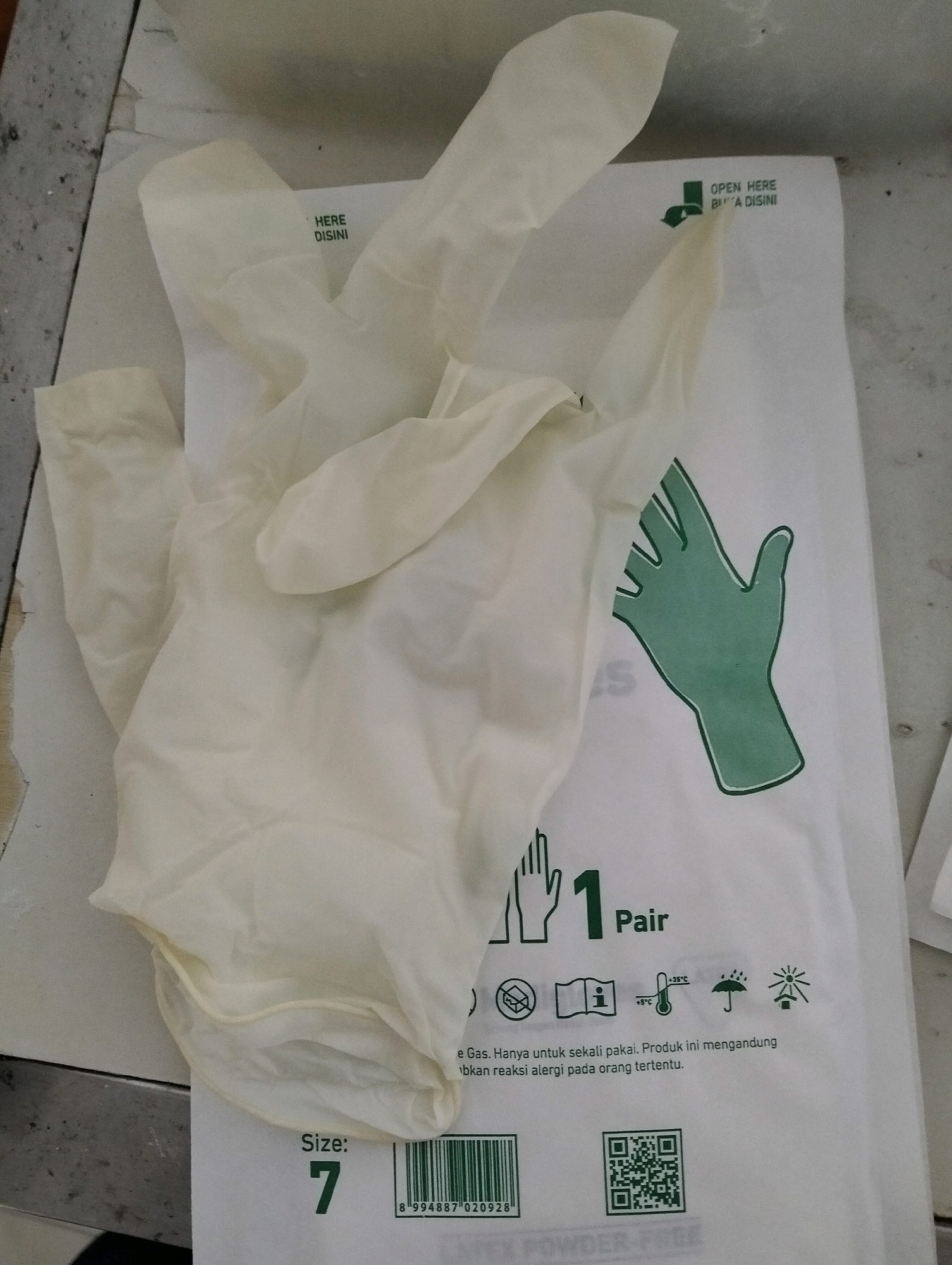 A pair of white latex gloves is laid out on top of a white paper packaging with green text and graphics. The packaging has text indicating "OPEN HERE" and "HERE DISINI", a green outline of a hand, "1 Pair", and several pictograms representing storage conditions and usage. Below this, the text "Size: 7" is visible, along with a barcode and a QR code. The image appears to be taken indoors, likely on a flat surface that could be a table or counter, with some debris visible on the left side. The overall impression is that of a product package for disposable gloves, presented in a neutral, sterile setting. The location is identified as Malang, Indonesia, although no specific landmarks or contextual clues in the image confirm this.