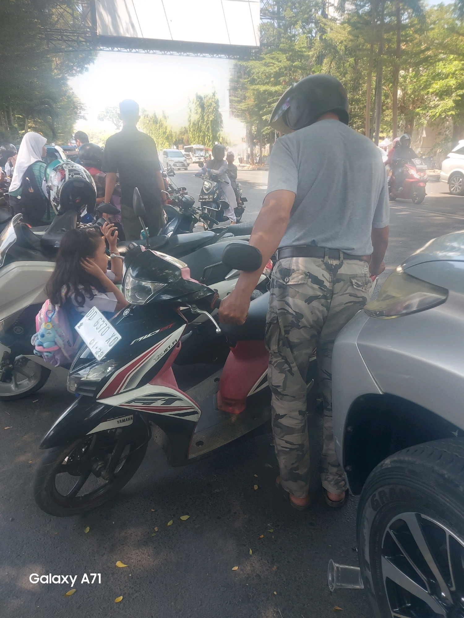 In the city of Makassar, Indonesia, on a bright, sunny day, a group of people are gathered around parked motorcycles on a paved road. A man wearing a grey t-shirt, a black helmet, and camouflage pants stands near a silver car, his back to the camera. In front of him, a child with long, dark hair sits on a white and pink Yamaha scooter, wearing a backpack with a Stitch plush toy. The scooter has a license plate that reads "DD 6370 XO 01.29". Other motorcycles and people are visible in the background, suggesting a busy street or gathering. Trees and a structure, possibly a large billboard or awning, are in the background, indicating an urban setting. The overall scene depicts a moment of everyday life in Makassar.