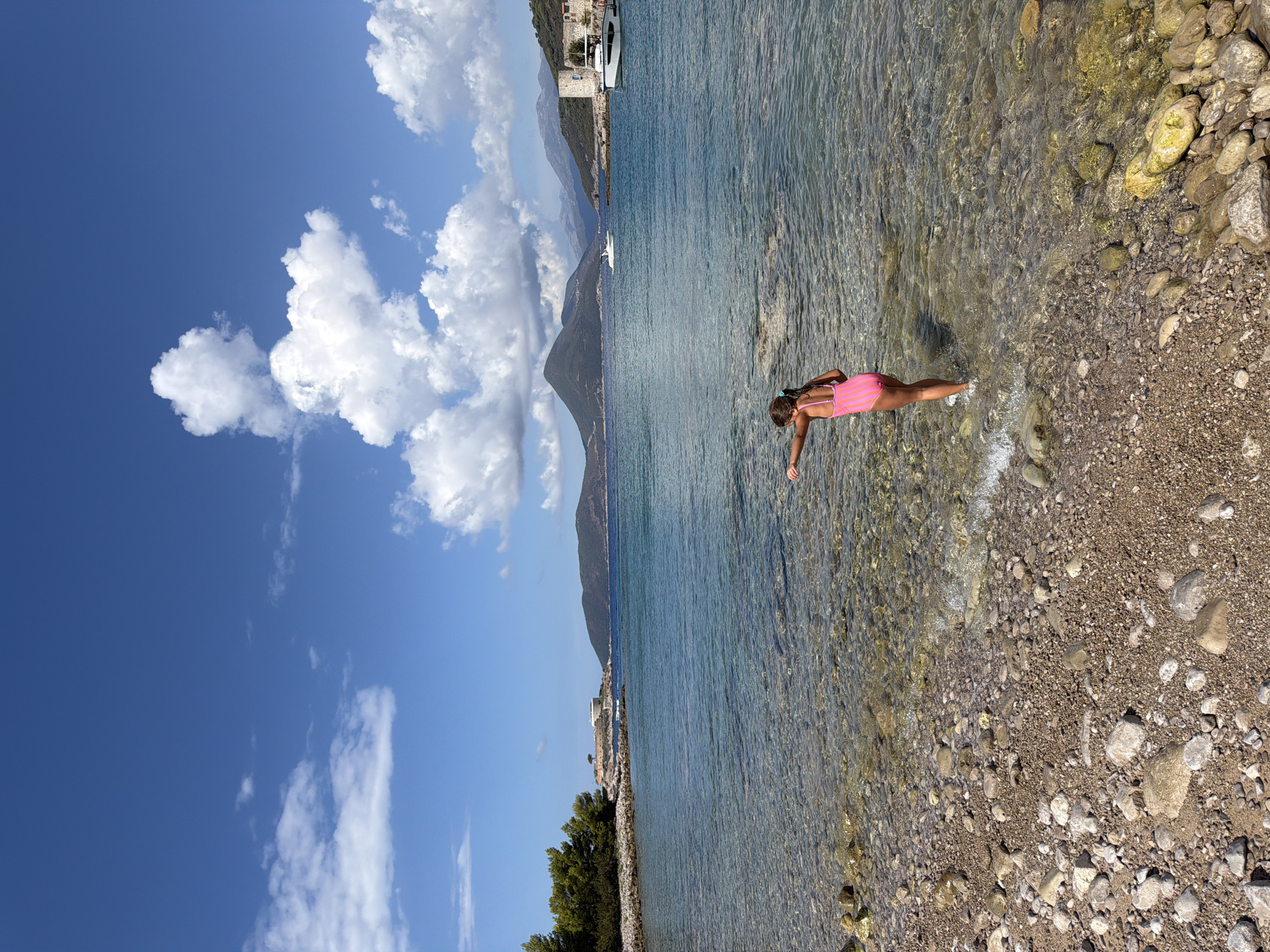 This media file depicts a sunny day at the coast of Luštica, Montenegro. The scene is dominated by a vibrant blue sky filled with fluffy white clouds. A young girl, wearing a pink and white striped swimsuit, is standing in the shallow, clear water of the Adriatic Sea. The water is calm, with small waves gently lapping at the pebble beach. The seabed is visible through the transparent water, revealing a mixture of sand and stones.
In the background, the coastline features lush green trees and a few buildings, suggesting a small settlement or resort. Further out at sea, a white boat is visible, and beyond that, a range of majestic mountains rises against the horizon, their peaks partially obscured by clouds. The overall impression is one of a peaceful and idyllic summer day at the beach, with a sense of relaxation and enjoyment. The sunlight indicates it's daytime, likely mid-morning or afternoon, given the brightness and clear skies. There is no visible text in the media file.