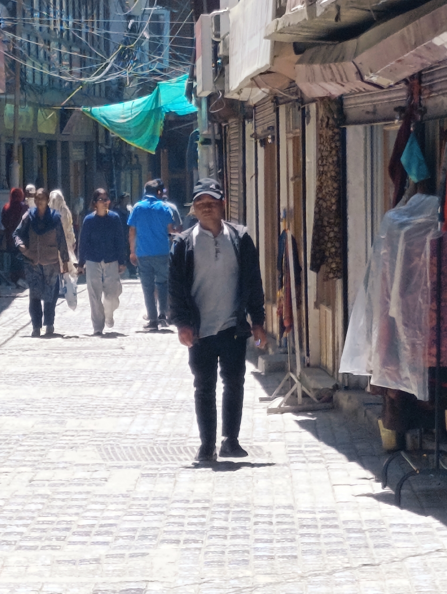 A medium shot captures a street scene in Leh, India, under bright daylight. In the foreground, a man walks towards the camera, wearing a black baseball cap, a grey t-shirt, a dark jacket, and black pants. To his left, on the sidewalk, are several other pedestrians. A woman in a brown jacket and patterned pants walks away from the camera, her left hand holding a white bag. Next to her, a woman in a dark blue sweater and light grey pants also walks away. Behind them, a man in a bright blue t-shirt and jeans walks away from the camera, his back visible. Further in the background, other figures are partially visible. The street is paved with a light-colored, brick-like pattern. The buildings lining the street are multi-story structures with shops on the ground floor. A turquoise tarp is stretched across the street overhead, creating shade. Wires are strung haphazardly across the street between buildings. Storefronts display goods, some covered in clear plastic. The overall impression is of a bustling marketplace street in Leh.