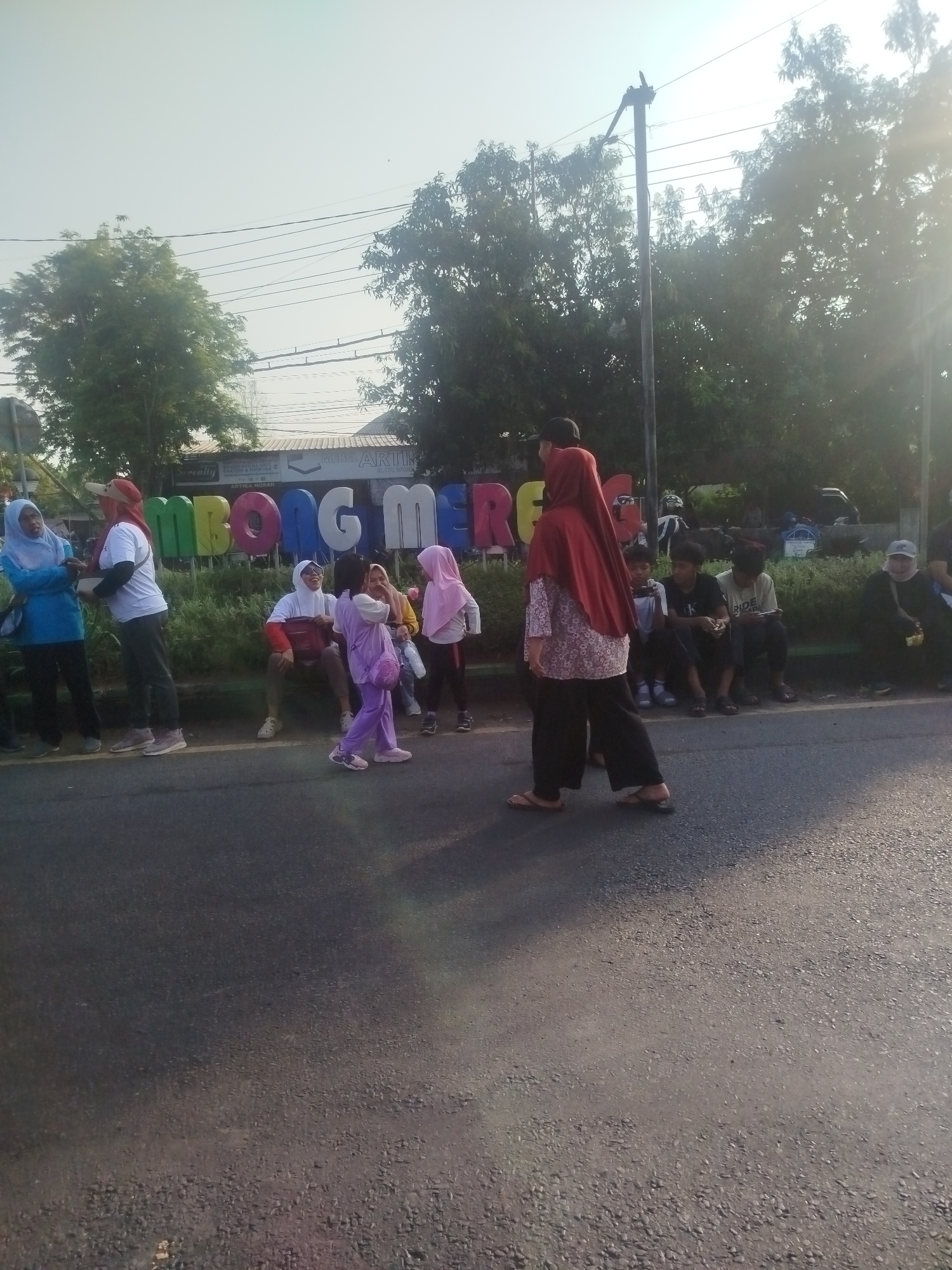 The image shows a street scene in Burneh, Indonesia. A group of people are gathered near a large colorful sign that spells out "MBONG MERCHANDISE". The sign is located in front of a building with a white roof. The people in the foreground include women and children, most of whom are wearing hijabs. Some of the women are standing and talking, while others are sitting on the ground. There are also some men and boys in the background, some of whom appear to be sitting on the ground as well. The street is paved with asphalt, and there are trees and power lines visible in the background. The lighting suggests it is daytime, with the sun casting shadows and creating lens flare in the upper right corner of the image.