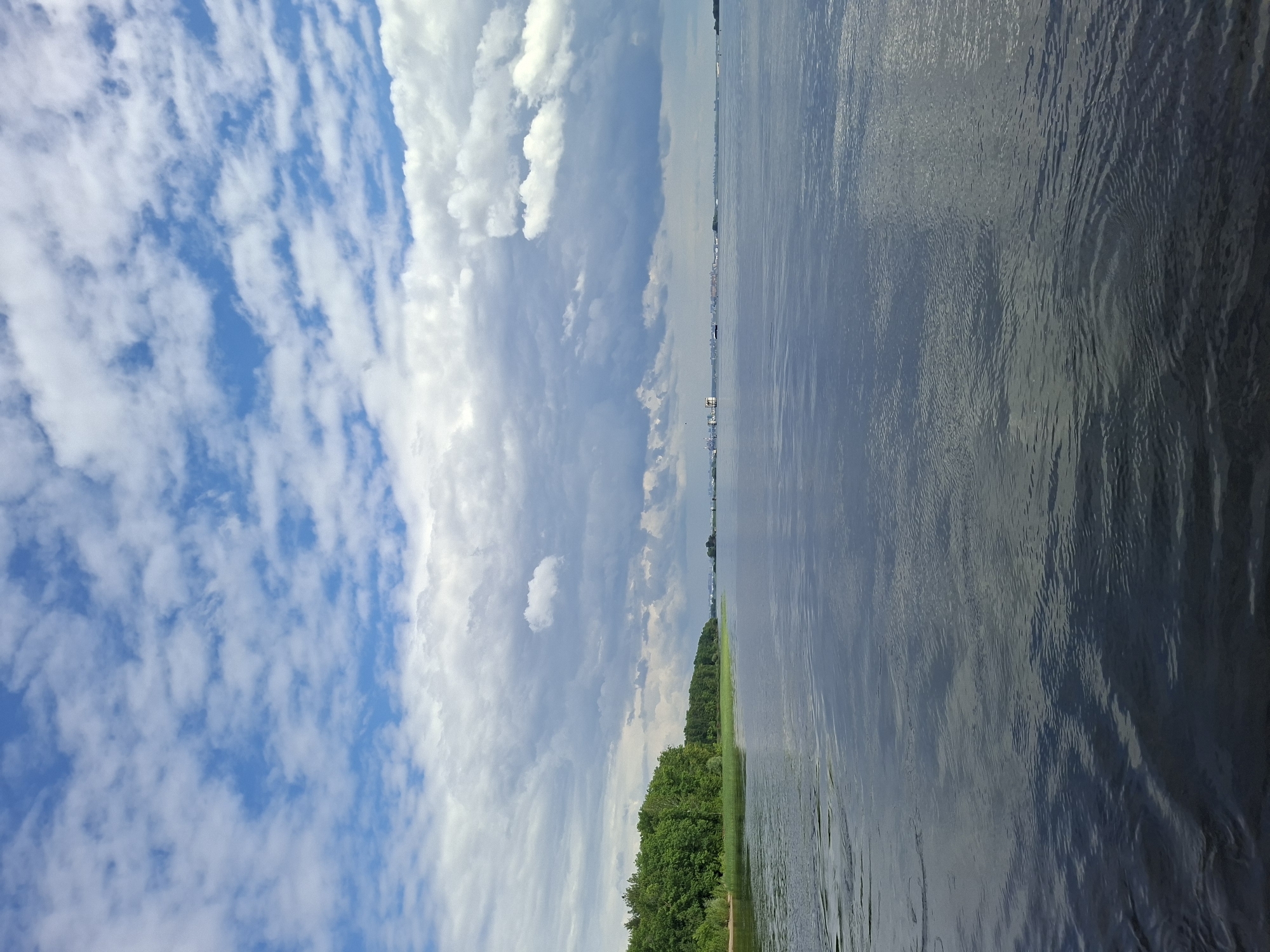 A wide, calm river occupies the majority of the foreground and midground, reflecting the sky and surrounding landscape. The water's surface is slightly rippled, creating a textured sheen. Along the left bank, a dense line of lush green trees forms a natural barrier, bordering a sandy shore. In the distance, across the river, a hazy cityscape is visible, with low-rise buildings and what appear to be industrial structures or port facilities. The sky is a dynamic mix of bright blue patches and dramatic white and gray clouds, with sunlight filtering through, casting soft illumination on the scene. The overall impression is one of a serene natural landscape meeting the edge of a populated area. This scene is consistent with a view along the Volga River near Kazan, Russia, a major waterway that flows through the city. No people or animals are visible in the image.