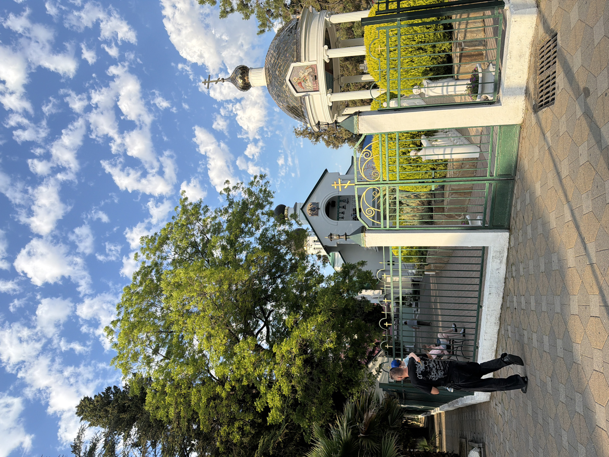 The image depicts an exterior view of a religious complex, likely a church or monastery, situated in Sochi, Russia. In the foreground, a man walks away from the camera, carrying a child on his hip, both dressed in casual attire. The man is wearing a black t-shirt with a graphic design and dark pants, while the child wears a blue hat. To the left of the man, a woman sits on a bench, with another person partially visible behind her. The pathway in front of them is paved with hexagonal tiles.
The scene is bordered by a green metal fence with decorative elements and crosses. Behind the fence, lush green bushes are visible, some of which are manicured into hedges. A white, multi-columned rotunda with a golden dome topped by a cross stands prominently to the right. Adjacent to it, a white church building with a dark, onion-shaped dome and a cross can be seen. Architectural details like windows, bells, and religious iconography are visible on the buildings.
The environment suggests a sunny day with a bright blue sky dotted with fluffy white clouds. Tall trees, including a large deciduous tree with vibrant green foliage and a tall conifer, frame the scene. The overall impression is one of a peaceful, well-maintained religious site.
