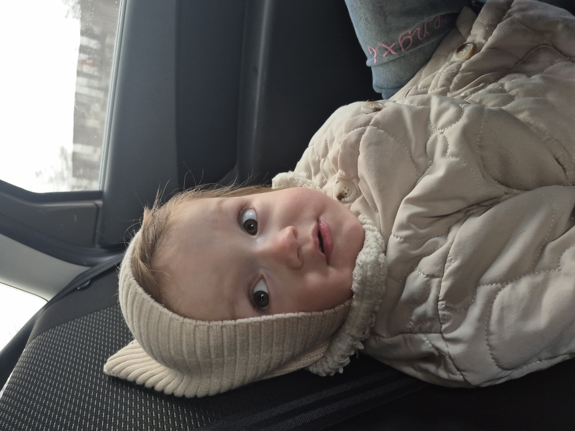 A close-up shot shows a baby lying on a black car seat, seemingly secured with a seatbelt across their torso. The baby wears a light beige quilted jacket with buttons and a matching knitted head wrap. A partial view of a window reveals a blurry outdoor scene with what appears to be snow-covered ground. Part of a blue jeans leg is seen on the top right of the image with pink writing. The location is Chaikovsky, Russia.