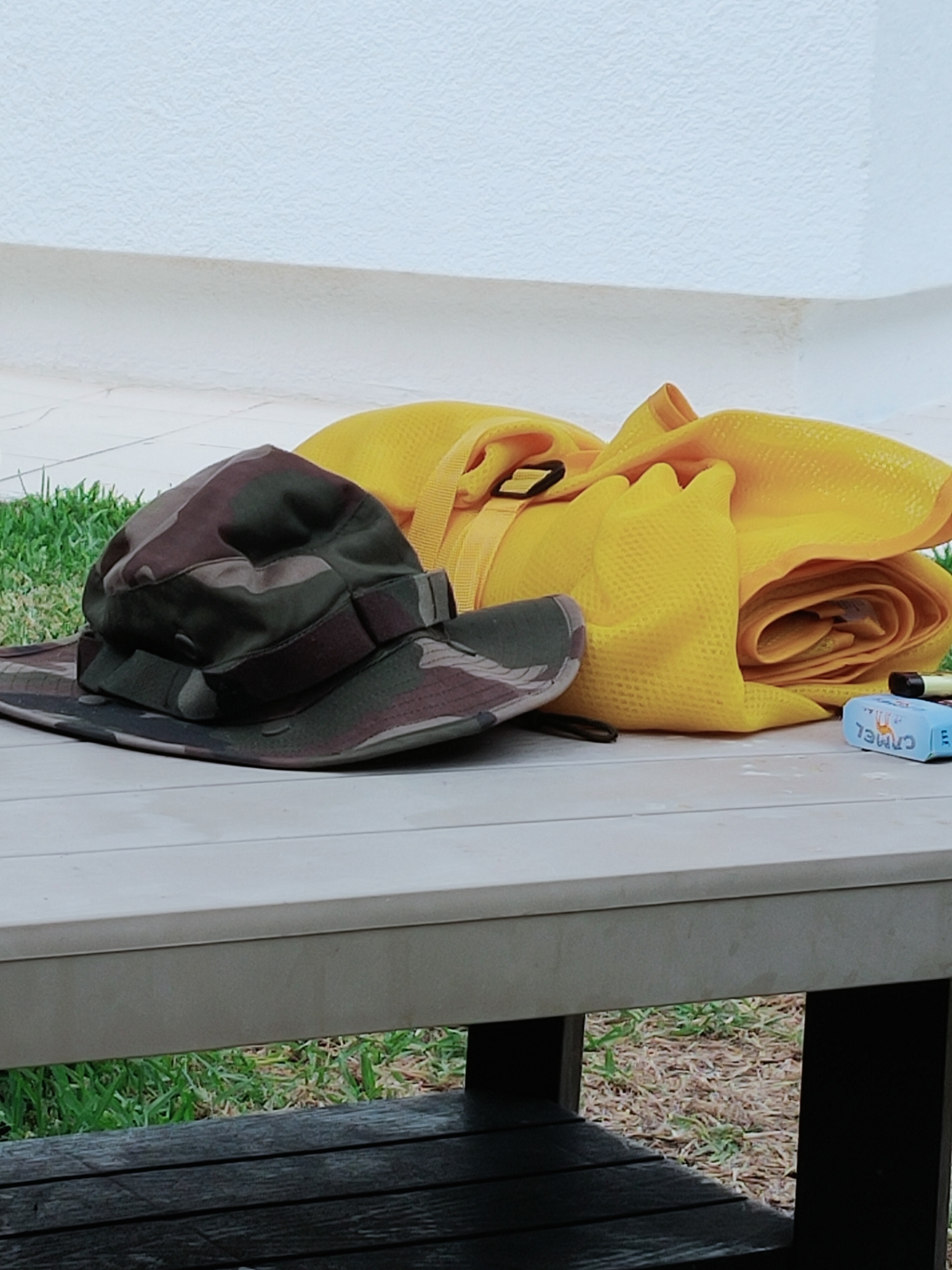 In the city of Mrezga, Tunisia, on a weathered, light-colored wooden surface, a camouflage-patterned boonie hat rests to the left. To its right, a bright yellow fabric, possibly a rolled-up mat or towel, is positioned. Next to the yellow fabric, a small blue rectangular object with white lettering, possibly a pack of cigarettes, is visible. The background consists of a textured white wall above a strip of green grass. The scene is viewed from a slightly elevated perspective.