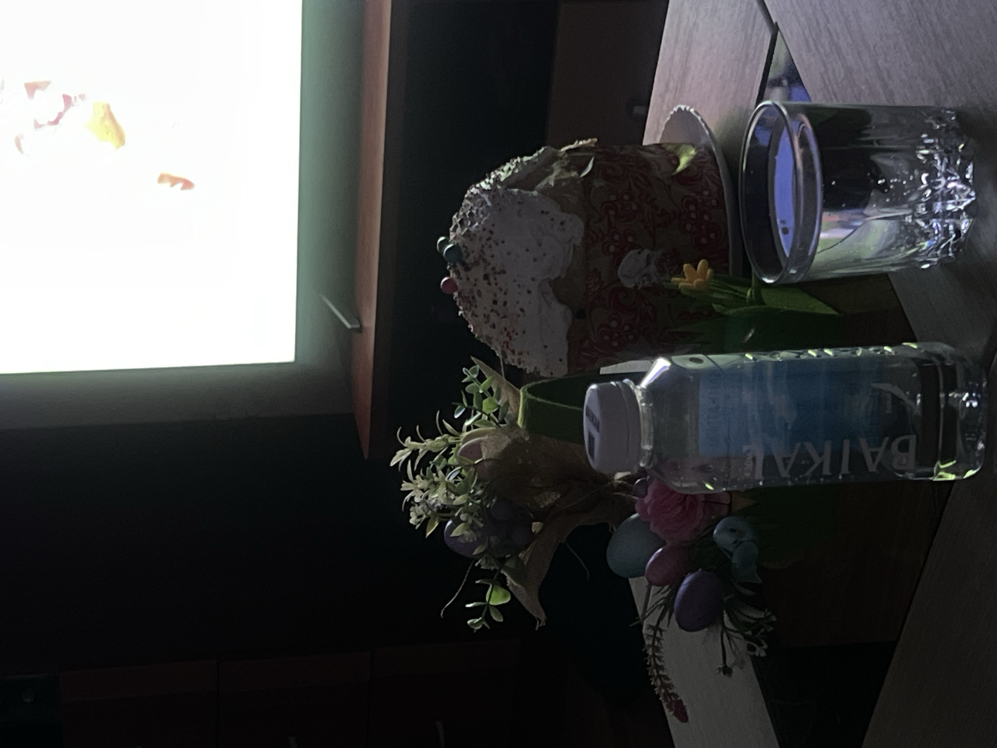 A still life sits on a table in a dim room, adjacent to a bright television screen. The composition includes a traditional Easter cake with white frosting and colorful sprinkles. Accompanying the cake is a decorative flower arrangement with artificial flowers, including Easter eggs and a single, small sunflower. A clear glass cup with water and a plastic bottle of Baikal water, labeled in what appears to be Russian Cyrillic script, are also visible on the table.
Given the presence of "Baikal" water and the traditional Easter cake, it's plausible the scene is in Russia, but the exact location within Ust-Ilimsk, Russia, cannot be determined from the image alone. The scene is domestic and still.