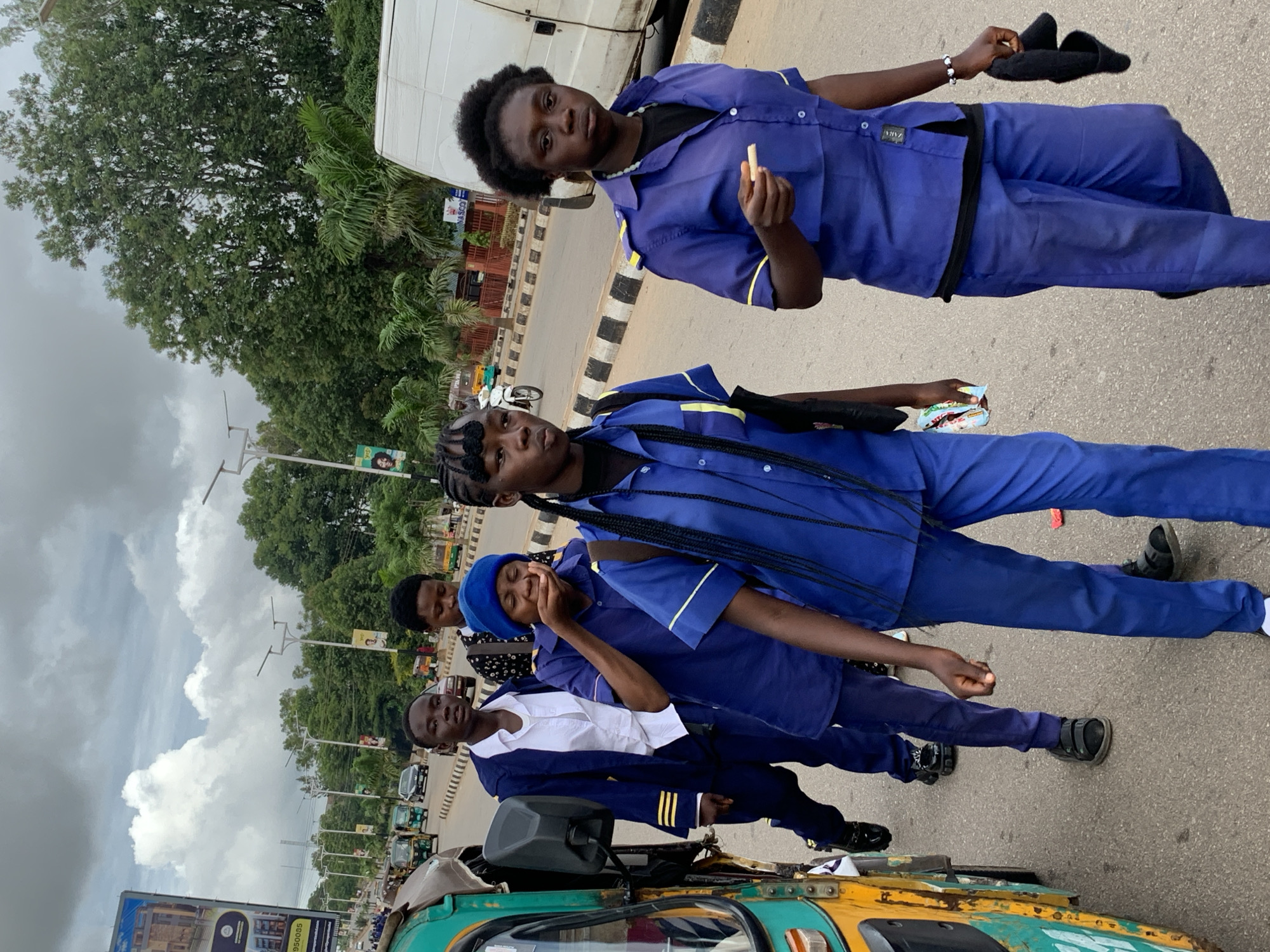 The image shows a group of young people walking on a paved road in an urban environment, likely Barakin Lerawi, Nigeria. The individuals are dressed in blue uniforms, with some wearing blue shirts with yellow trim and navy blue trousers. One person in the foreground is holding a white object, possibly food or a pen, in their hand. To the left of the group, a colorful auto-rickshaw is visible, partially obscuring some of the individuals. The road has a distinct black and white striped curb. In the background, there are trees, streetlights, other vehicles including an auto-rickshaw and a white van, and buildings. The sky is cloudy. The overall scene depicts a street in a city with a sense of movement and daily activity.