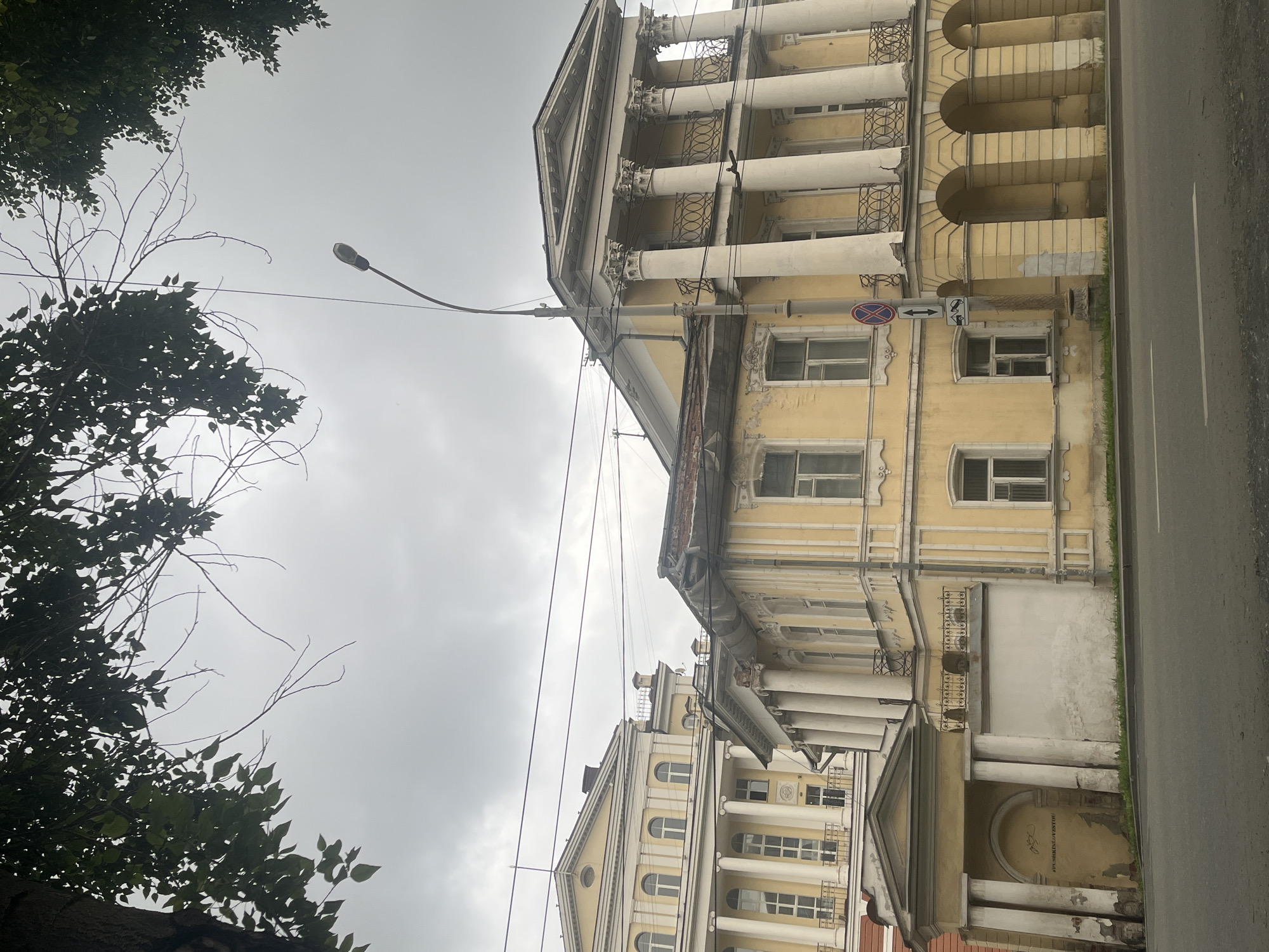 The image shows an exterior view of classical architecture on a cloudy day. In the foreground, a grey asphalt road runs horizontally across the bottom of the frame, with white lane markings visible. Behind the road, a low white wall with a small archway and columns is present on the left. Adjacent to it, a light yellow building features a series of arched openings supported by columns, with a balcony above. This building connects to a taller, also light yellow building with multiple windows and a prominent colonnade on its facade. The taller building is partially obscured by the foreground structure. Overhead, tree branches with green leaves frame the top of the image, and a lamppost is visible extending upwards towards the overcast sky. Several power lines are strung across the sky. The overall scene suggests an urban setting with historical architecture.
