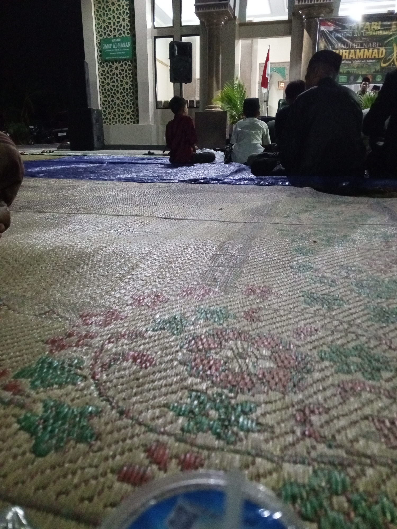 The image shows a nighttime scene outside the Jami Al-Hasan mosque in Lemahbang, Indonesia. Several people are seated on mats on the ground, facing the mosque. A banner is visible behind them, with text that appears to be related to a Maulid Nabi Muhammad celebration. The mosque has a distinctive green and white patterned facade. A red and white flag is also visible inside the mosque entrance. Two large speakers are placed on the ground to the left of the mosque entrance. The foreground is dominated by a textured mat with a floral pattern.
