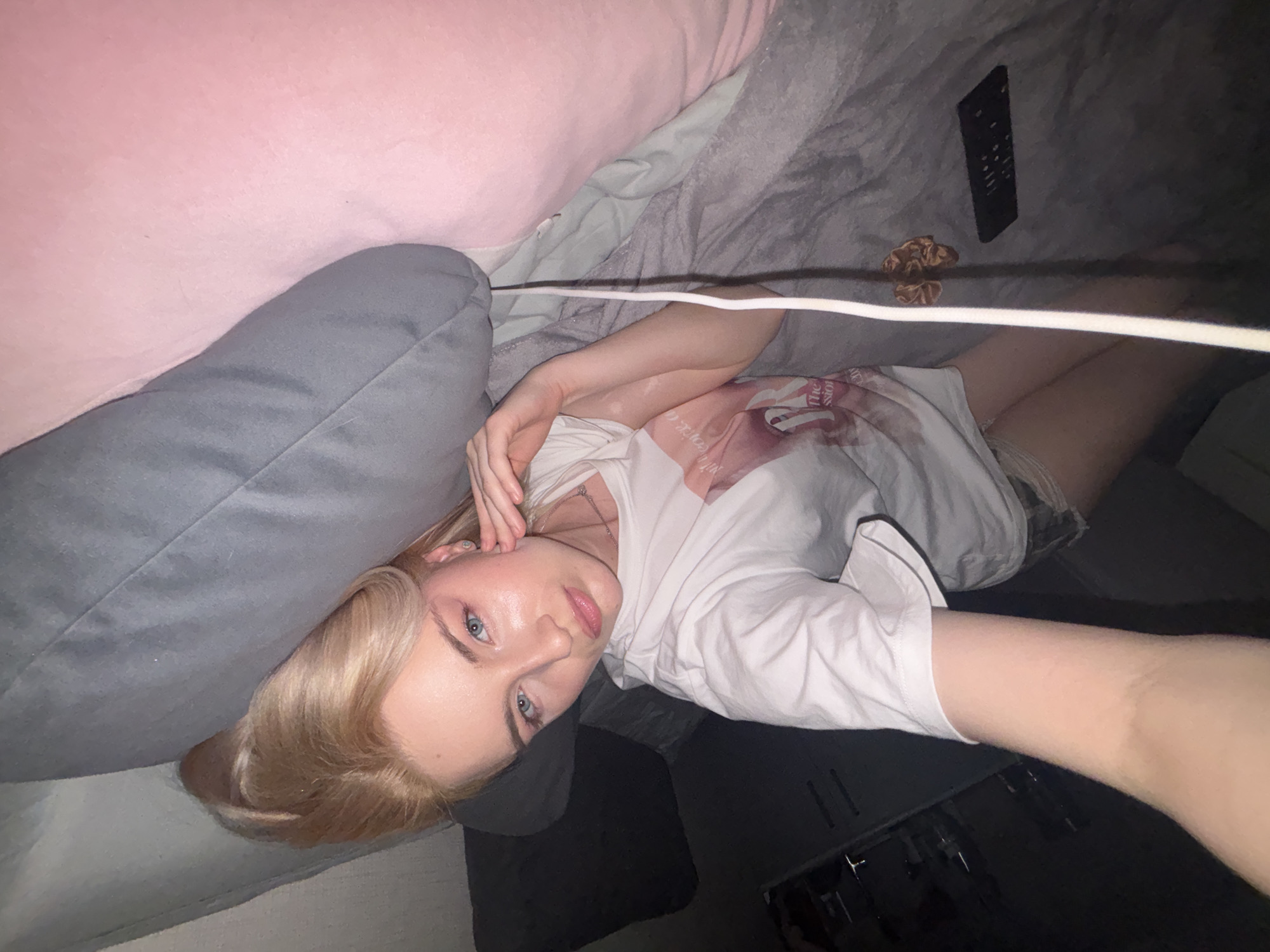 The image depicts a young individual, likely female, situated indoors in what appears to be a bedroom or living area within the city of Unknown, Russia. The individual has light blonde hair styled with significant volume, blue eyes, and light-toned skin, exhibiting apparent hypopigmented patches on the left arm. She is wearing a white short-sleeved t-shirt with a partial graphic showing text including "The Passion" and a word starting with "Sion," along with light-colored shorts or undergarments visible beneath the shirt. A small stud earring is present in her left ear. Her left hand is raised, with fingers lightly touching her left cheek and ear. She is lying down, extending her right arm towards the camera, presumably holding the device used to capture the image. A large dark gray pillow is positioned behind and partially above her head. To her right, a large pink, soft surface is visible, likely another pillow or bedding. A light gray blanket covers the lower portion of the scene. On this blanket, a white cable runs upwards, alongside a brown scrunchie and a black television remote control. The lighting is dim, suggesting an indoor setting with artificial illumination.