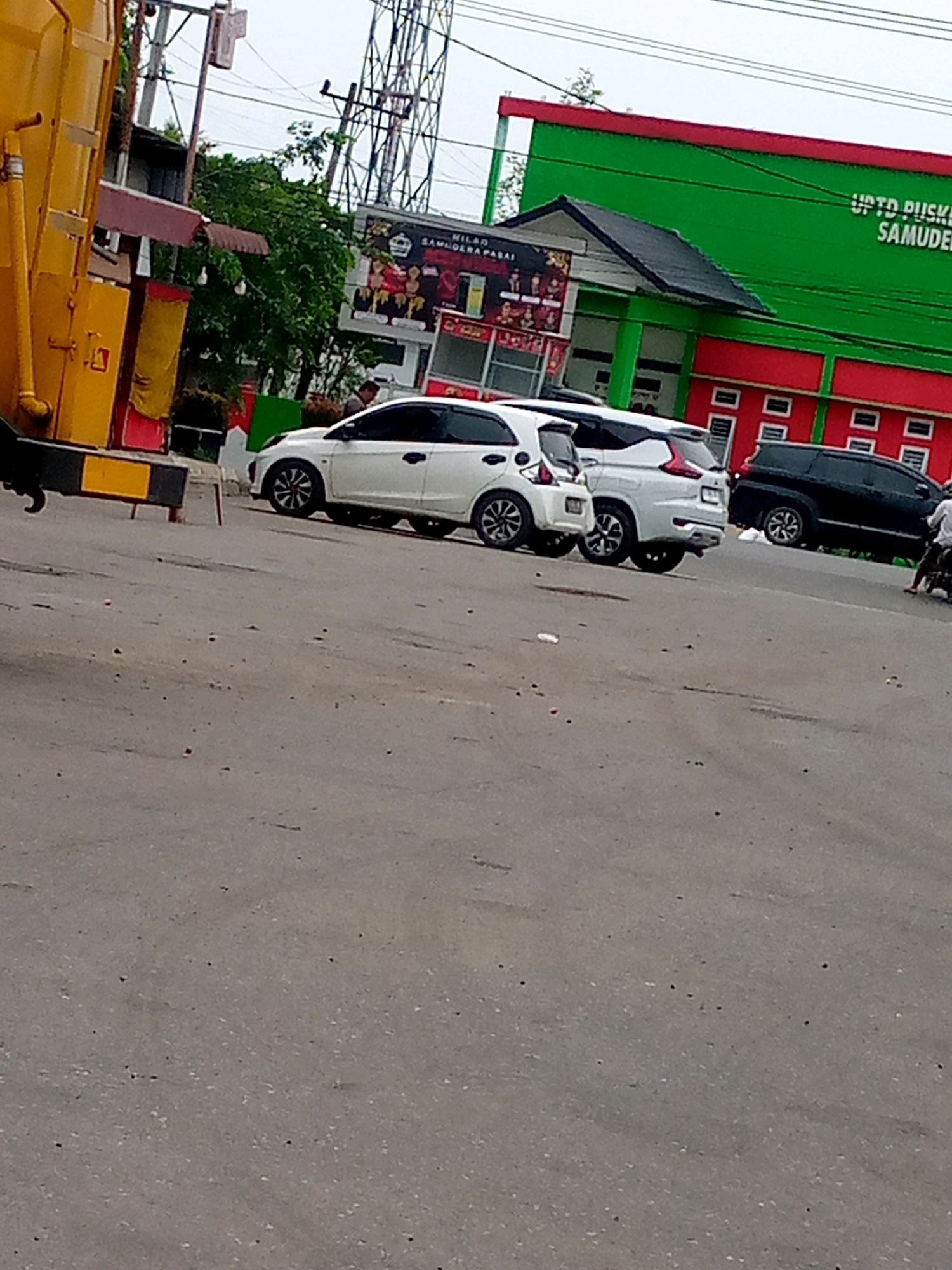 In Blangpeuria, Indonesia, a street scene unfolds with several vehicles present. A yellow and black striped traffic barrier stands on the left side of the frame. Adjacent to it is a large yellow structure, possibly part of a garbage truck. Parked on the asphalt road are two white cars, one slightly behind the other, and a white SUV. Further down the road, a black SUV is parked, and a person on a motorcycle is visible passing by. In the background, a green building with red trim and red window accents is prominent, displaying signage that reads "UPTD PUSKESMAS SAMUDERA." Trees and a cell tower are also visible in the background.