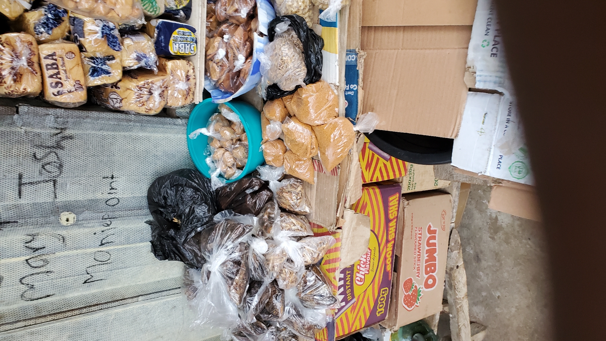 The image shows a roadside market stall in Jalingo, Nigeria. The stall is stocked with various food items, including bread, snacks, and what appear to be dried goods. Some of the items are packaged in plastic bags, while others are in boxes and bowls. There are handwritten labels on the wall behind the stall, including "Mary Joshua" and "Monief oint." The overall impression is one of a busy, informal market setting.