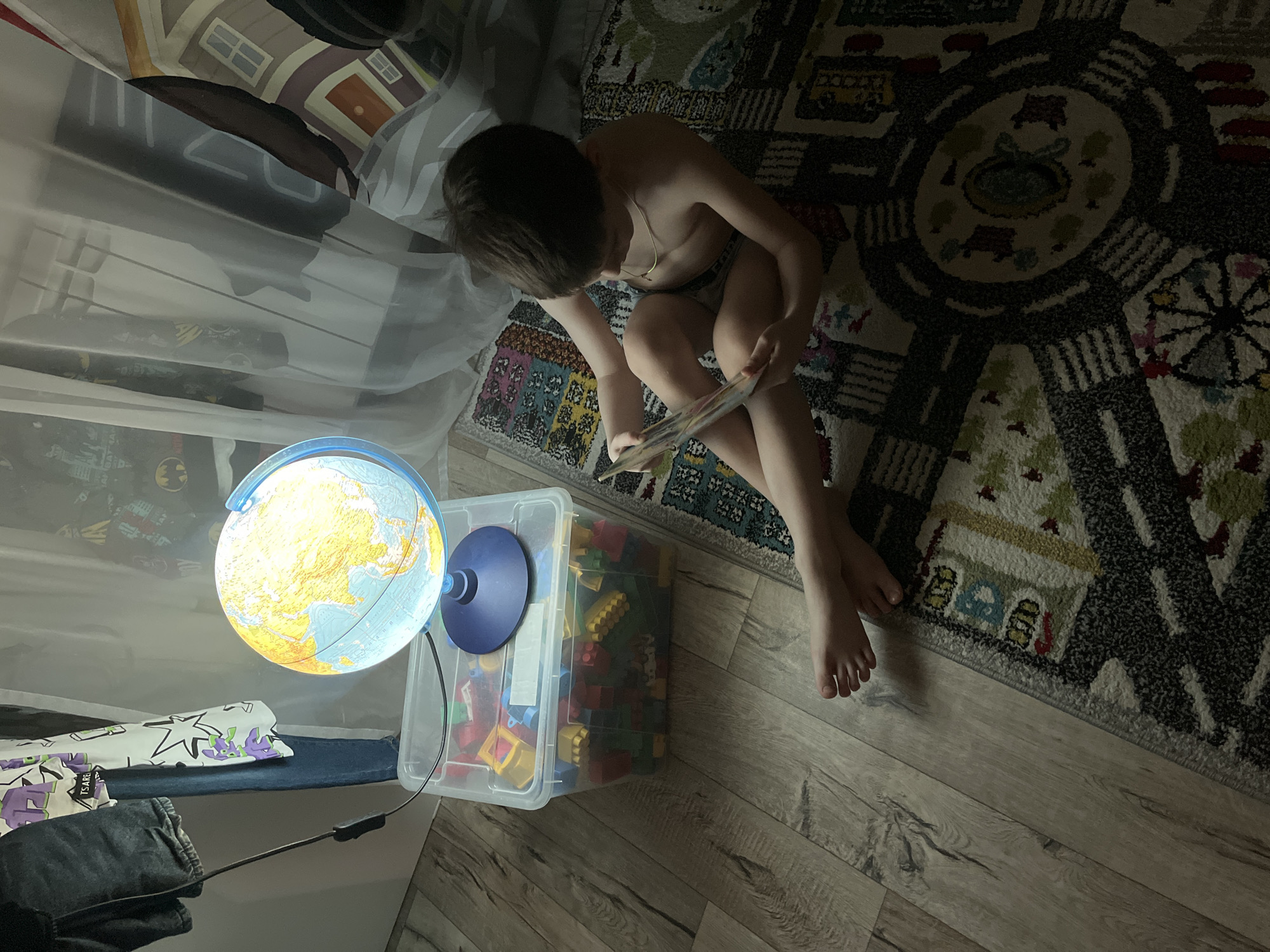 An indoor scene in Chaikovsky, Russia, depicts a dimly lit room with a single child present. The child, a bare-chested individual approximately 4-7 years old with dark hair and a thin gold-colored necklace, is seated cross-legged on a multi-colored rug with abstract patterns, potentially depicting a cityscape. The child is focused on reading a colorful book held open in both hands.
To the left of the child, an illuminated globe, predominantly yellow and light blue, rests on a dark blue circular base. This globe is positioned on the clear lid of a plastic storage bin filled with various colored building blocks (red, yellow, blue, green). A black power cord extends from the globe towards the left, passing over the bin.
The immediate floor surrounding the rug is light-colored wooden laminate. In the background, to the left, dark jeans and a white t-shirt with purple and green cartoon patterns are hanging, partially obscured by a sheer white curtain. Behind and through the curtain, elements of wall decoration or fabric are visible, including a dark pattern with white Batman symbols and a stylized illustration of a house with a brown roof and white walls. The primary light source in the scene appears to be the glowing globe.