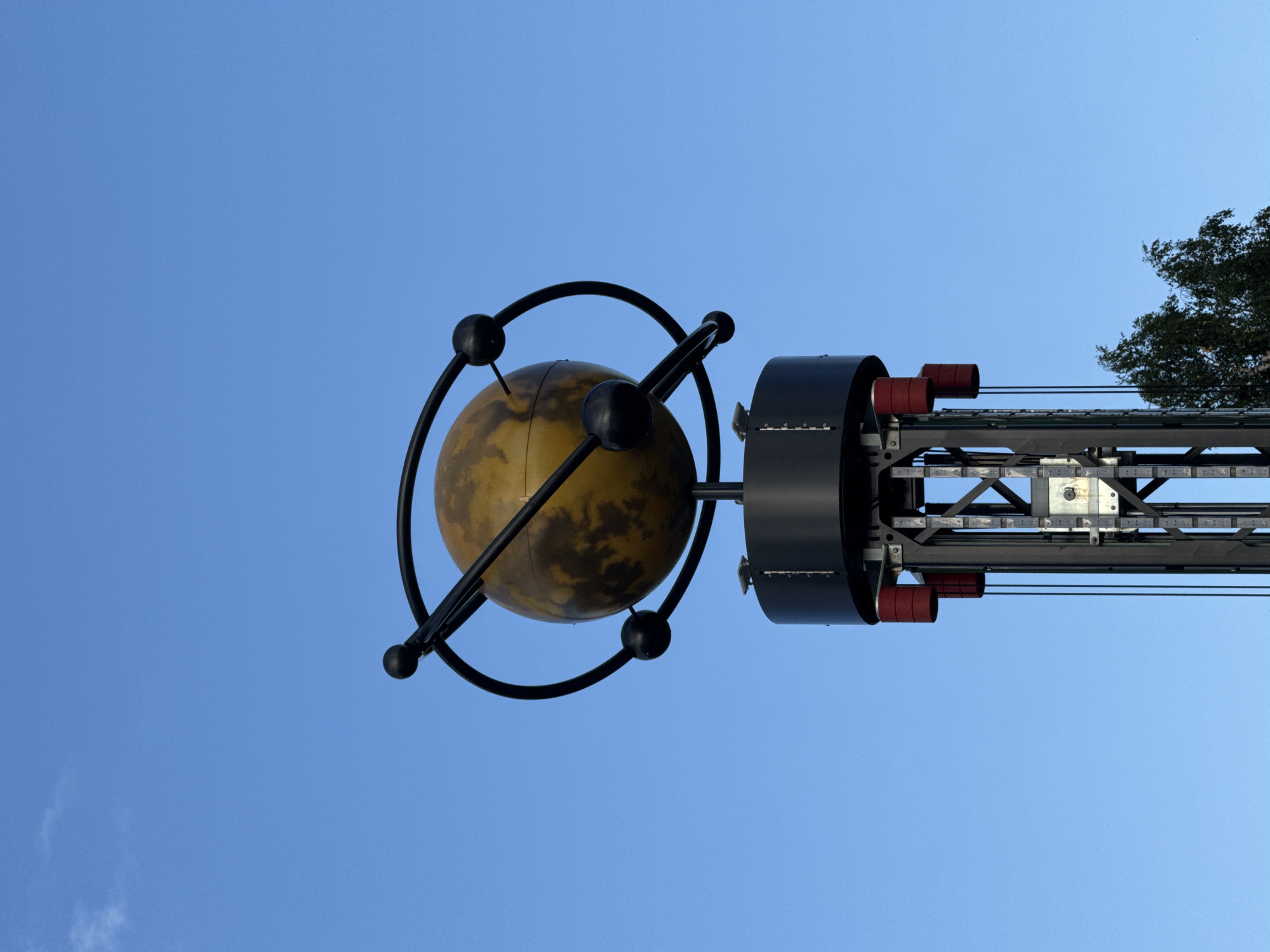 A low-angle, full-frame shot features a large, abstract globe sculpture mounted atop a dark, industrial-looking tower. The globe, a sphere of mottled yellow and brown, is encircled by a black, elliptical ring with three small black spheres attached at intervals, resembling an atomic model. The tower is constructed of dark metal with visible support beams and what appear to be red cylindrical components near the top. The background is a clear, vibrant blue sky with a single, wispy white cloud. A few green tree branches are visible at the bottom right of the frame. This sculpture is located in Moscow, Russia.