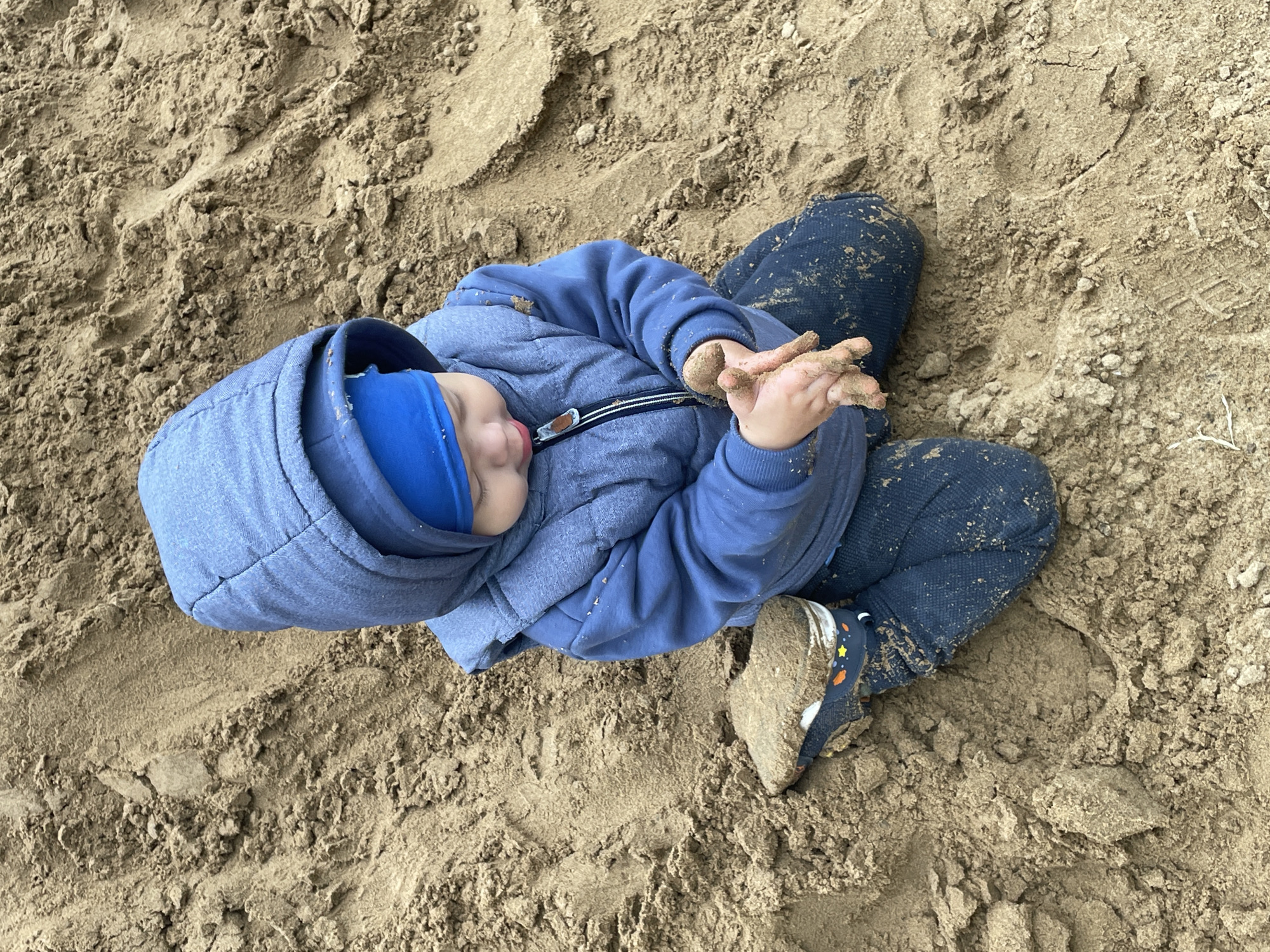 An overhead shot captures a young child engrossed in play on a sandy surface, likely a sandbox or a sandy play area in Perm, Russia. The child, appearing to be a toddler or preschooler, lies or sits partially reclined on the sand. They are dressed in warm attire suitable for outdoor play: a blue hooded jacket, a bright blue beanie, and dark blue pants, all speckled with sand. Their dark shoes feature a distinctive colorful star pattern and are also thoroughly coated in sand, particularly the left shoe visible in the frame.
The child's hands are a central focus, clasped together and generously covered in sand, suggesting they were actively digging or manipulating the sand. Their eyes are closed, and their mouth is slightly open, indicating a moment of deep concentration, peaceful rest, or perhaps even a sensory experience of the sand. The entire ground is composed of light brown, loose sand, showing various textures and disturbances from movement. The lighting suggests it is daytime, with soft, diffused light implying an overcast sky or shaded conditions, enhancing the focus on the child's interaction with their environment. No discernible text is visible on the child's clothing or in the sand. The scene conveys a sense of childhood innocence and joyful exploration of nature.