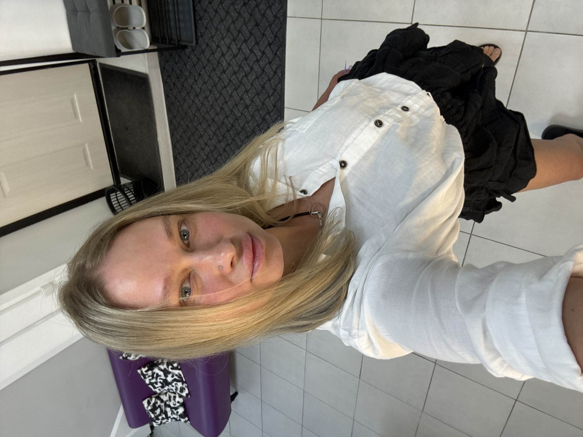 A selfie captures a Caucasian woman indoors, presumably in Perm, Russia, based on the provided location. She wears a white button-down shirt and a black skirt. Her blonde hair is loose. A purple chair with a black and white patterned cover is visible in the background. The floor is tiled. A door and a gray cushioned bench on a black frame are also in the scene.