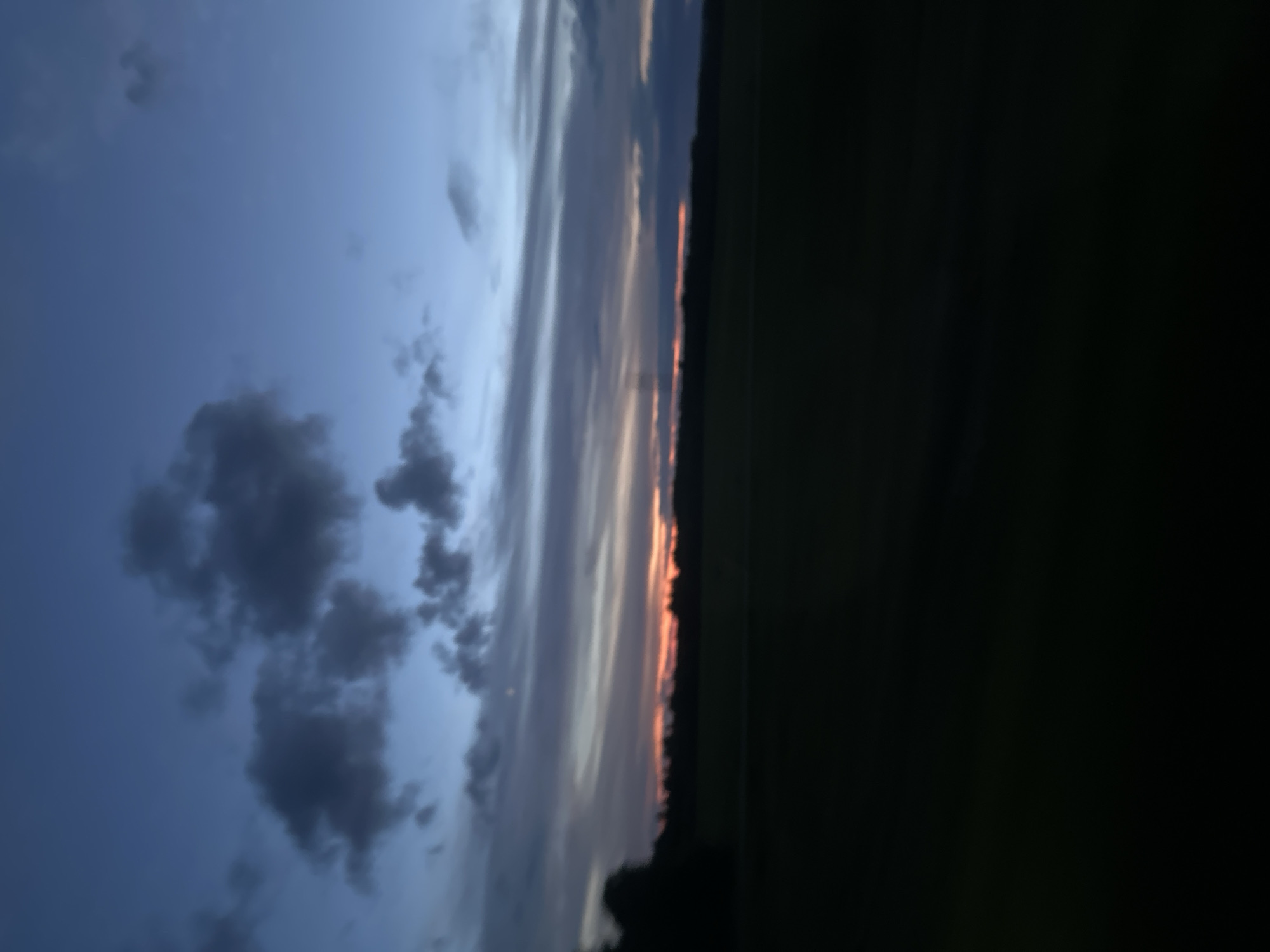 The image depicts a rural landscape at dusk in the vicinity of Грозаўскі сельскі Савет, Belarus. The sky is a deep blue, transitioning to lighter shades near the horizon, with streaks of orange and pink from the setting sun visible through scattered clouds. The clouds are dark and textured, adding depth to the sky. Below the sky, a dark silhouette of trees lines the horizon, suggesting a wooded area. In the foreground, a vast, dark expanse of what appears to be a field or open land occupies the lower half of the image, rendered almost entirely in shadow due to the low light conditions. There are no people or discernible objects visible in the frame, and the overall scene is one of natural, undisturbed twilight in a rural setting.