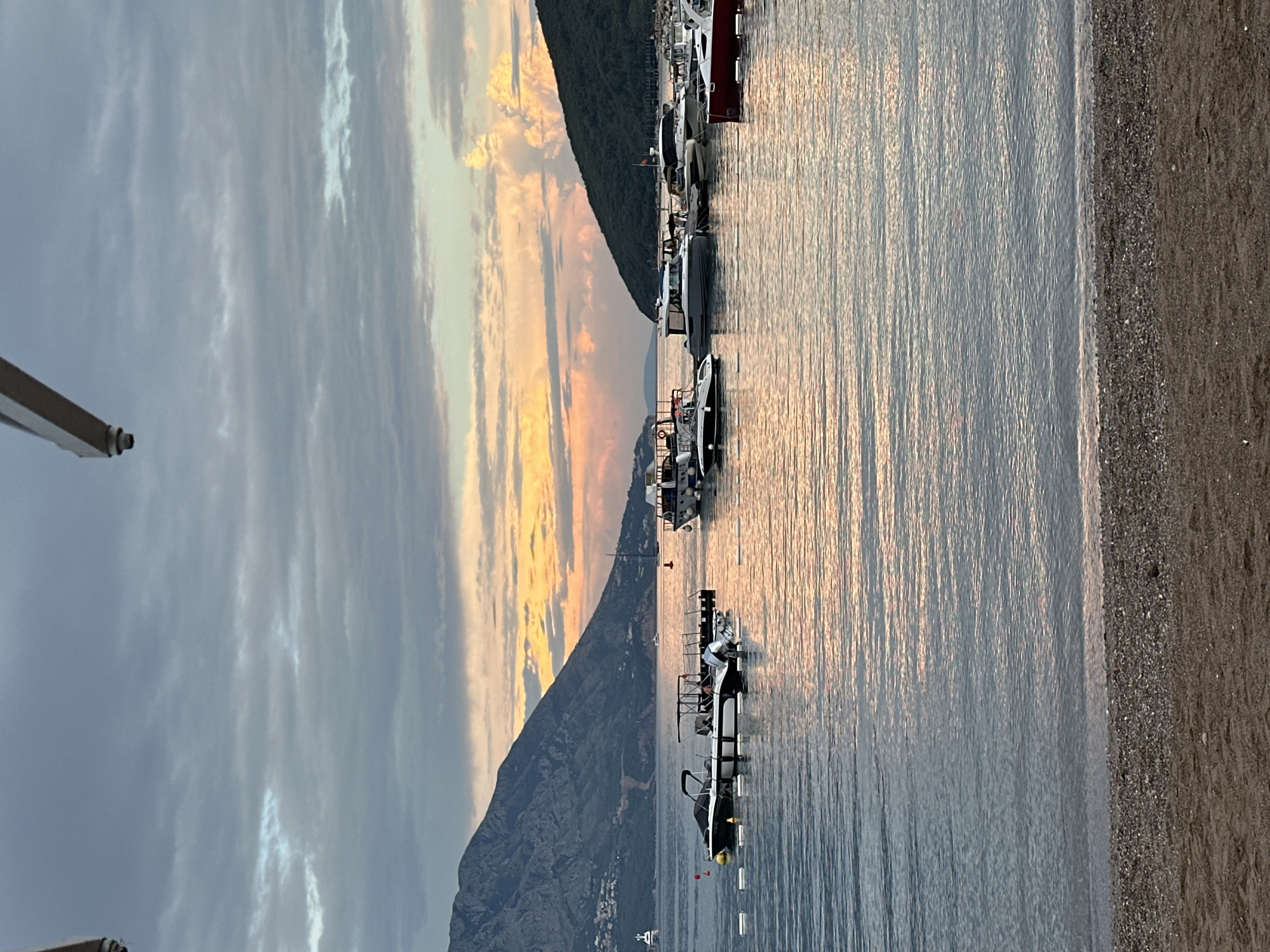 The image depicts a scenic bay at sunset, likely in Budva, Montenegro, given the characteristic mountainous coastline. The foreground shows a pebble beach transitioning into the calm, reflective water of the Adriatic Sea. Several boats are moored in the bay, ranging from smaller speedboats to larger yachts, with some anchored closer to the shore and others further out. The sky above is a canvas of soft, pastel colors, with clouds tinged with orange and pink hues from the setting sun, contrasting with the cooler, grey tones of the upper atmosphere. The backdrop is dominated by the lush, green hills and imposing mountains that are typical of the Montenegrin coast, with a hint of urban development visible on their slopes. A white structure, possibly part of a beach umbrella or awning, is partially visible in the top right corner of the frame. The overall atmosphere is serene and tranquil, capturing the beauty of a coastal evening.