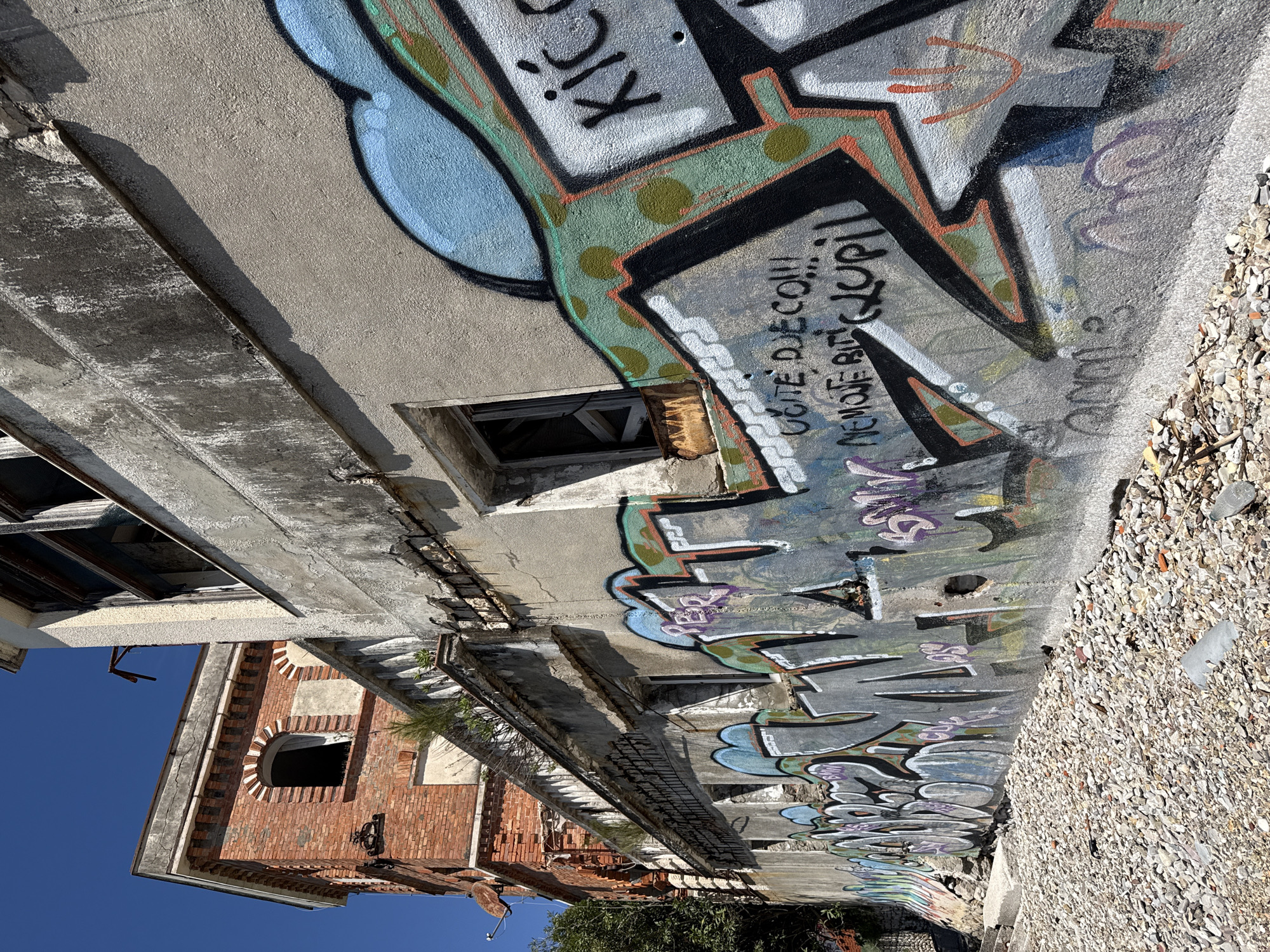 An exterior low-angle shot shows an abandoned building in Meljine, Montenegro. The building has exposed brick sections, deteriorating concrete, and multiple windows, some broken or missing. Graffiti covers the walls. The ground is a mix of rubble and debris. The sky is clear blue.